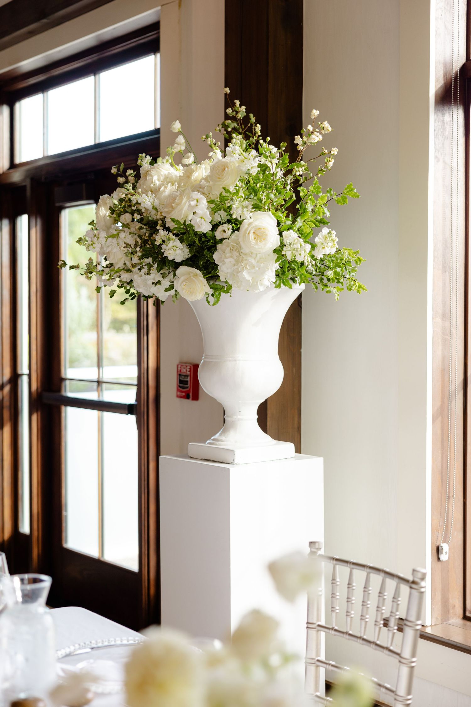 White floral arrangement in classic urn vase displayed on pedestal near window.