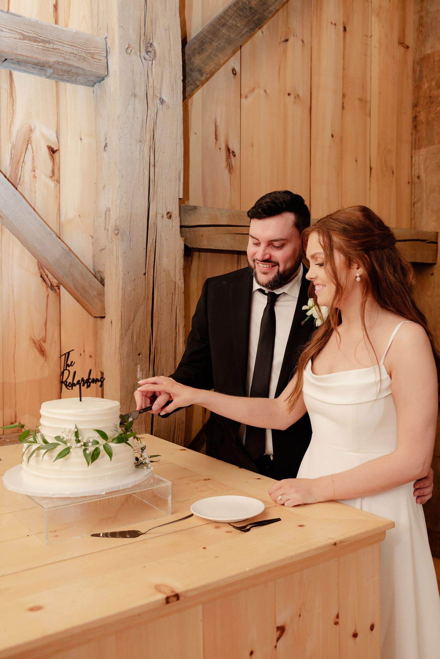 Newlyweds cut their white wedding cake together in a rustic barn setting with wooden walls.