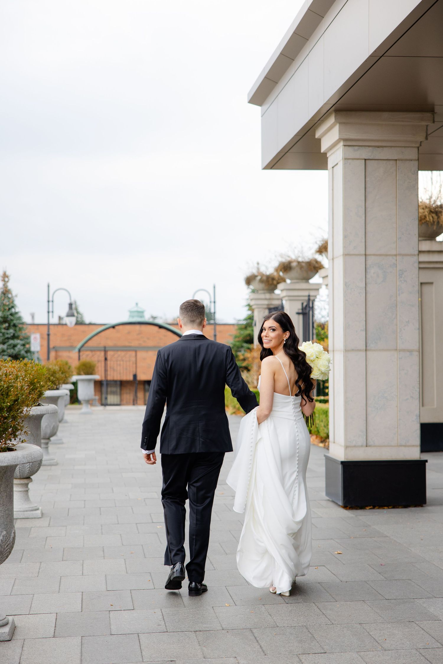 A couple in wedding attire stroll beneath classical columns on a stone terrace.