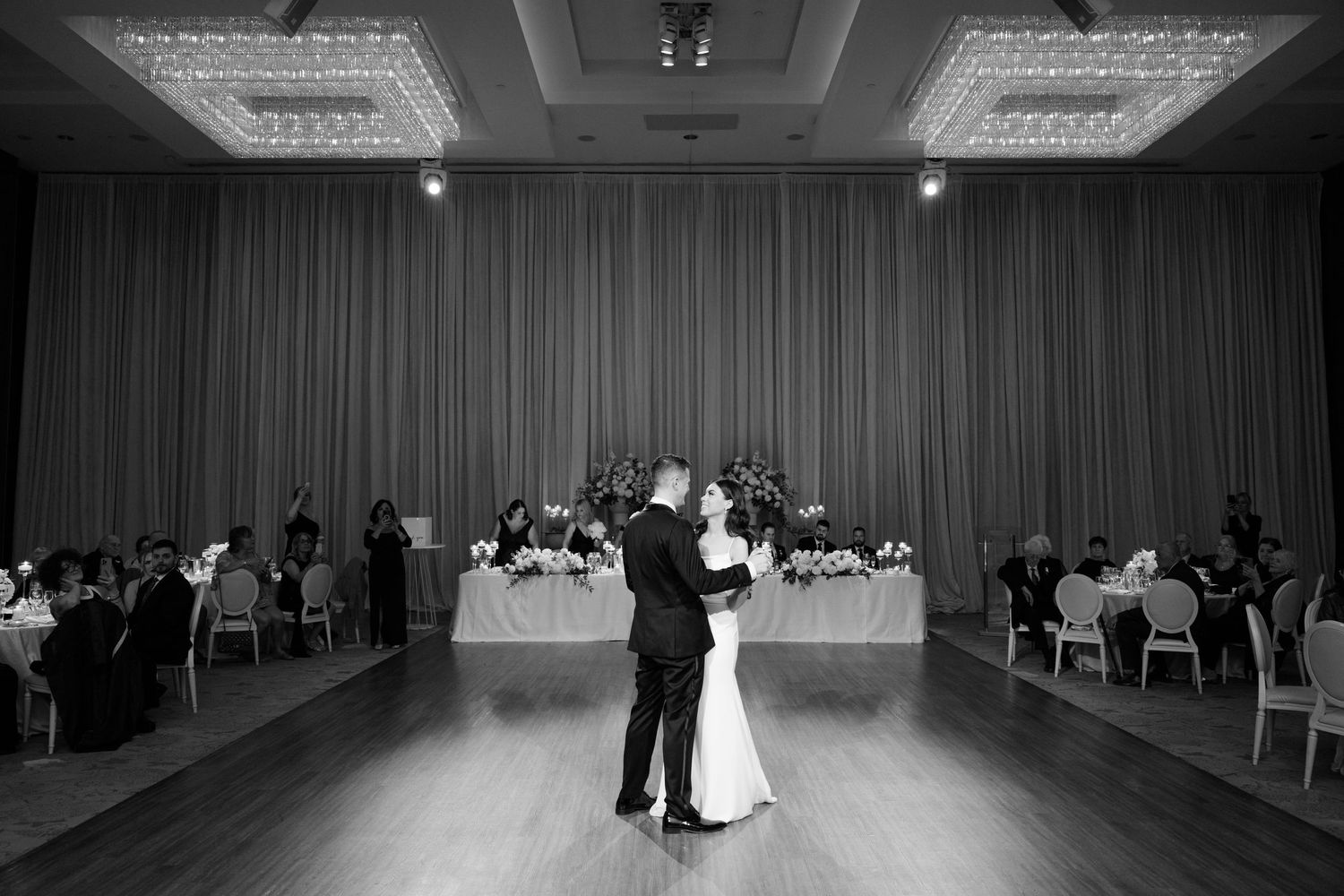 Black and white photo of newlyweds dancing alone on a ballroom floor with guests seated at tables.