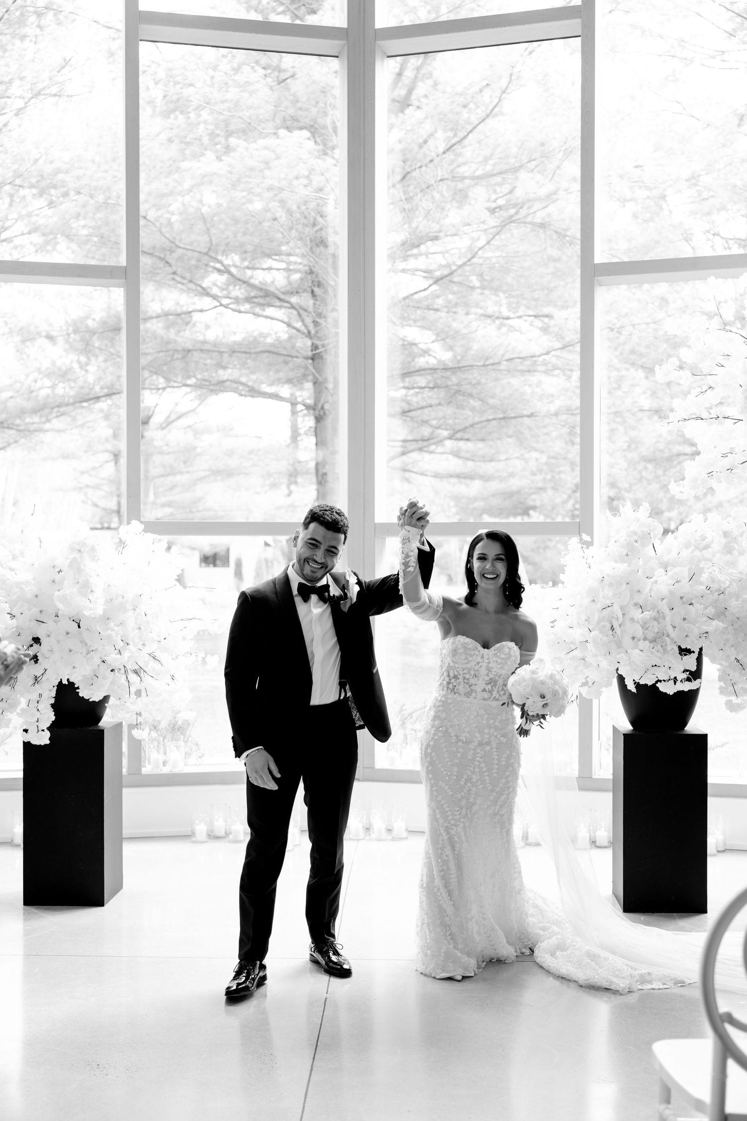 A black and white photo of newlyweds celebrating with raised hands in a light-filled venue.