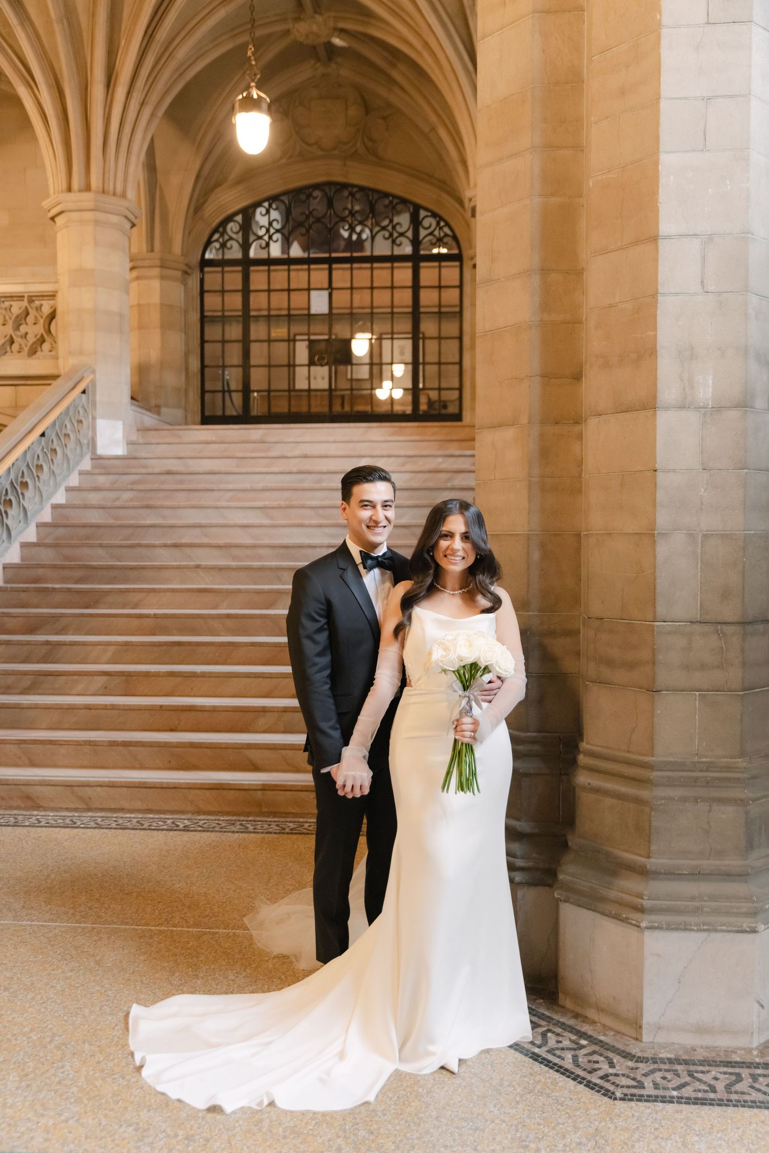 Elegant wedding couple poses on grand marble staircase in historic building with arched architecture.