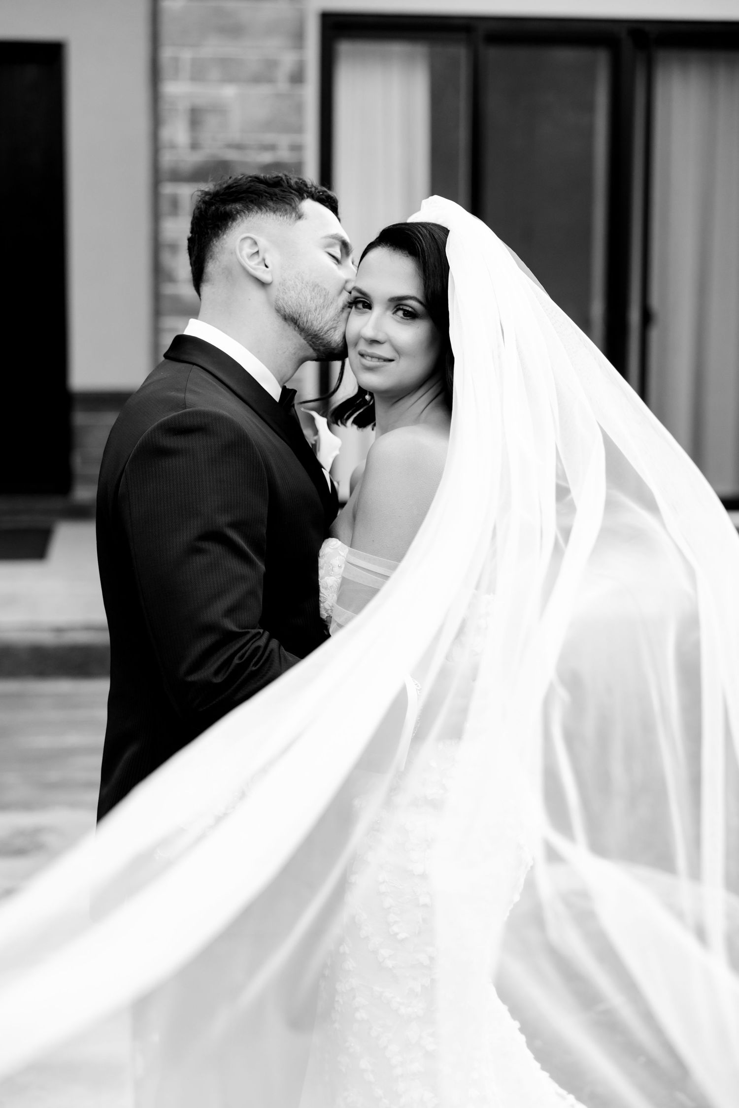 Black and white wedding portrait showcasing bride's elegant veil and couple's tender moment.