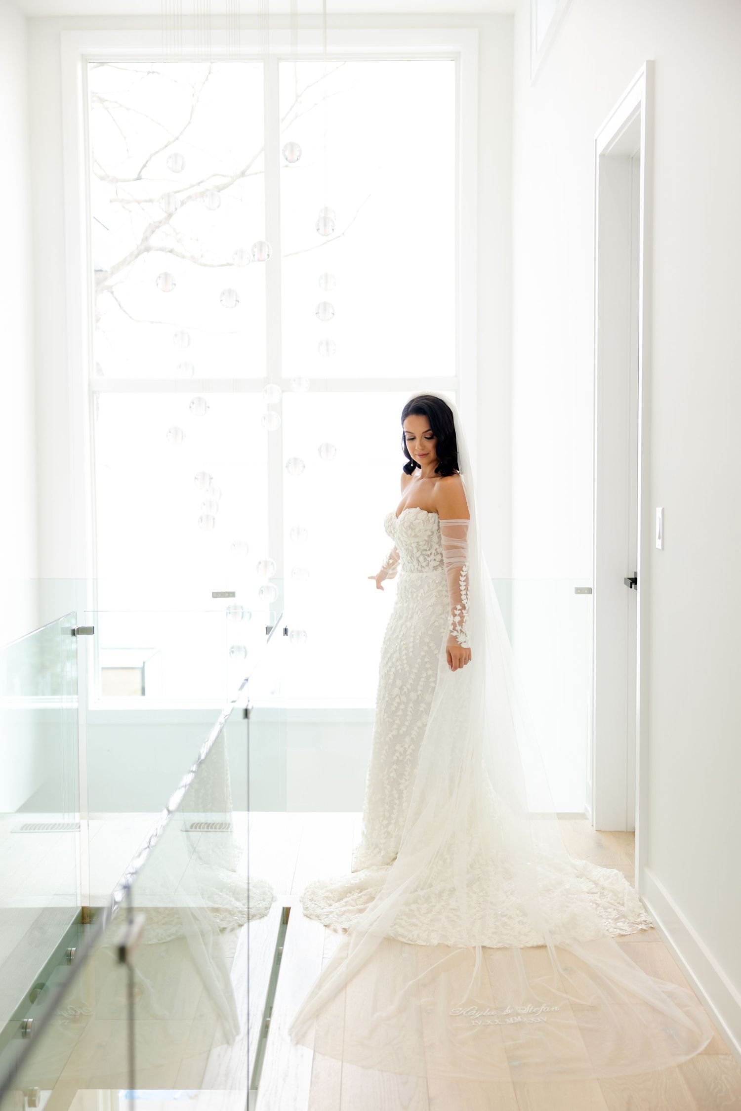 A bride poses in a modern bright hallway wearing a fitted off-shoulder wedding gown with delicate lace details.