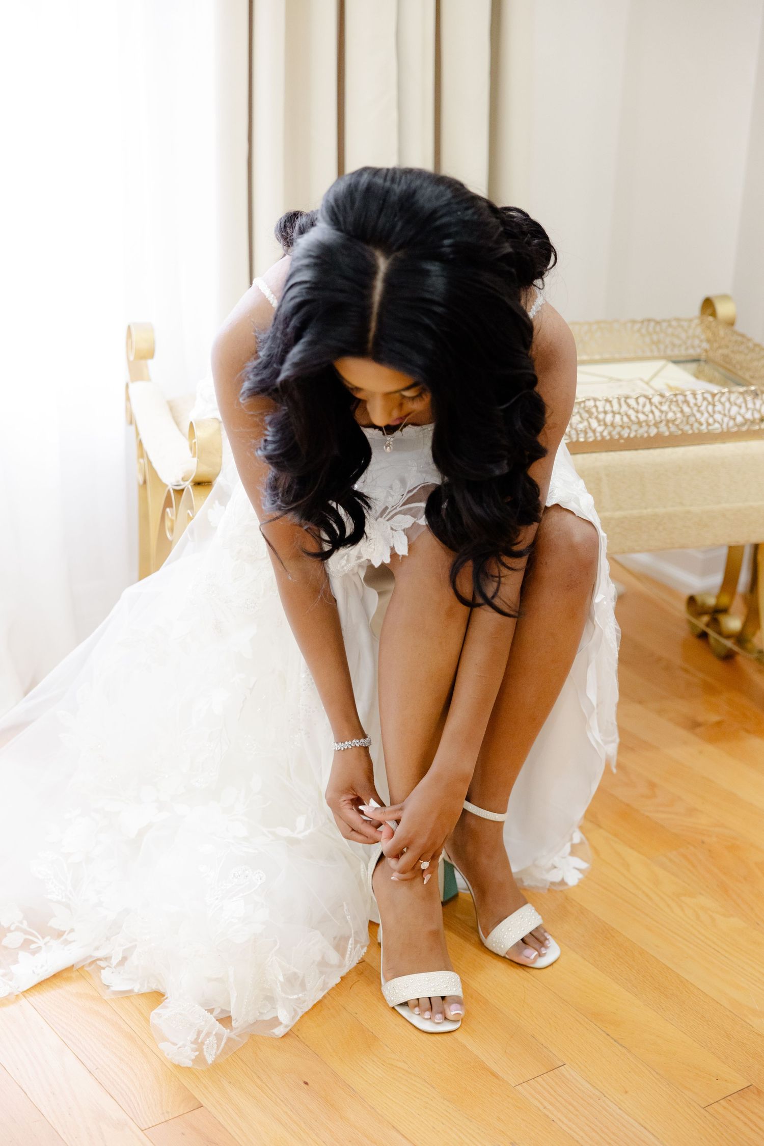 Bride in white dress fastens her strappy sandals while seated in a light-filled room.