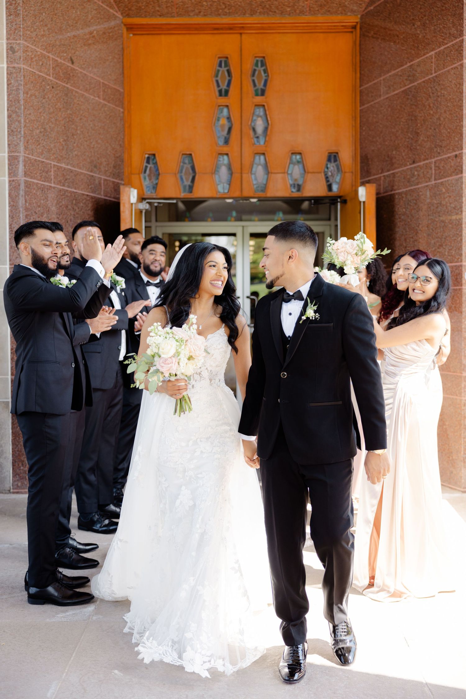 Newlyweds walk through church entrance as wedding party celebrates with raised bouquets.