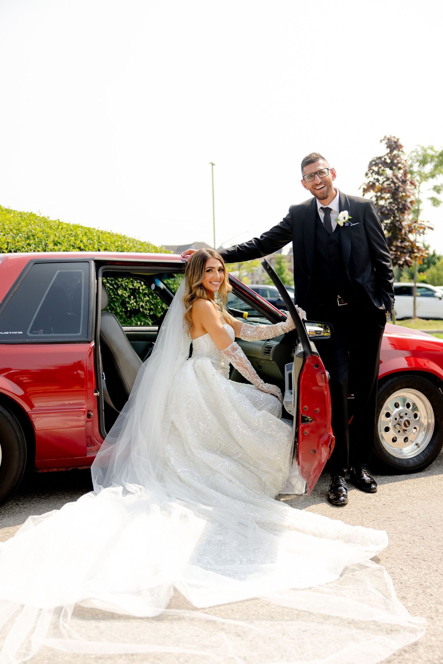 Wedding couple posing by a red car door with the bride's dress flowing beautifully.