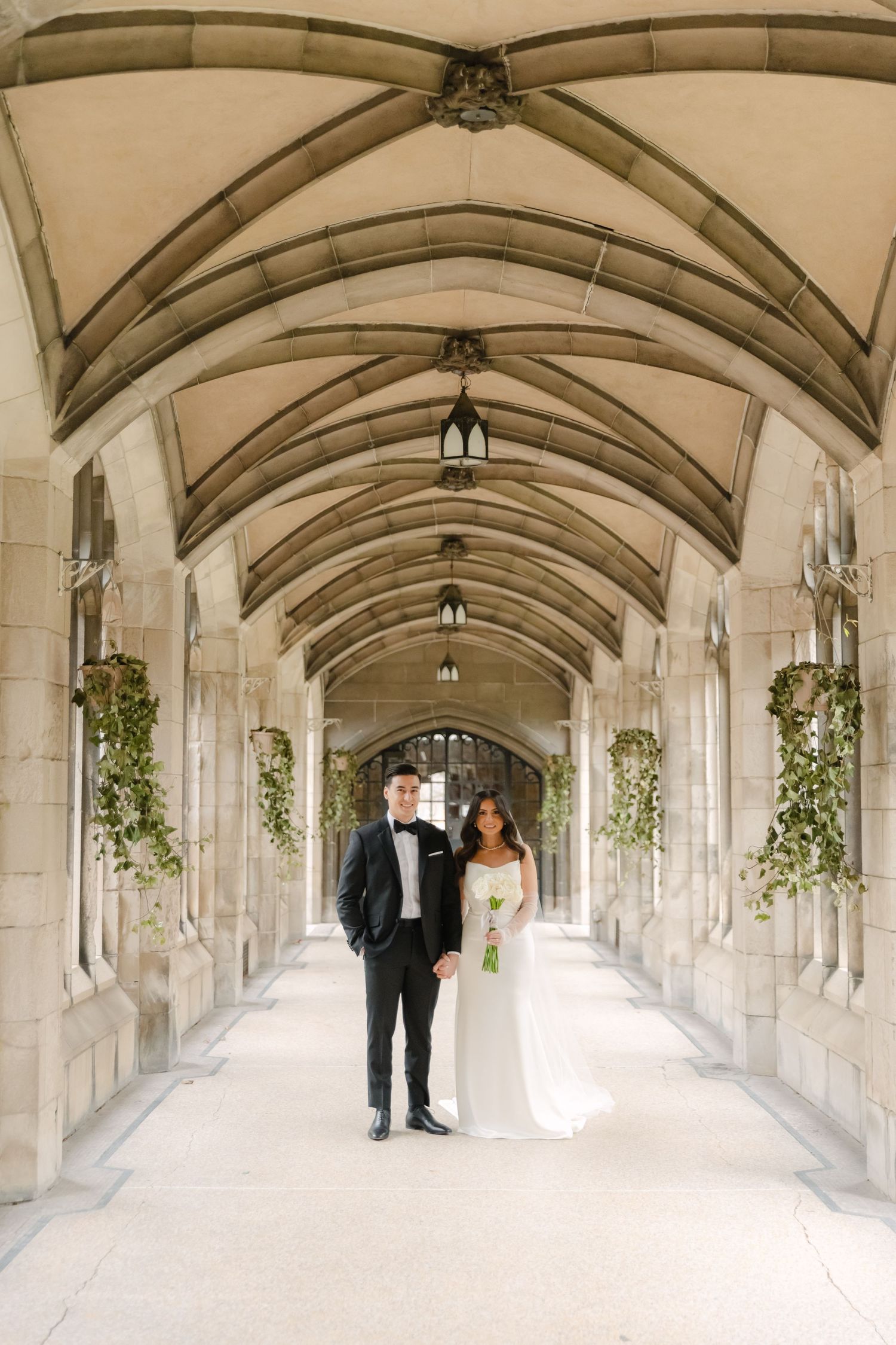 Wedding couple stands under vaulted stone ceiling decorated with vintage lanterns and greenery.