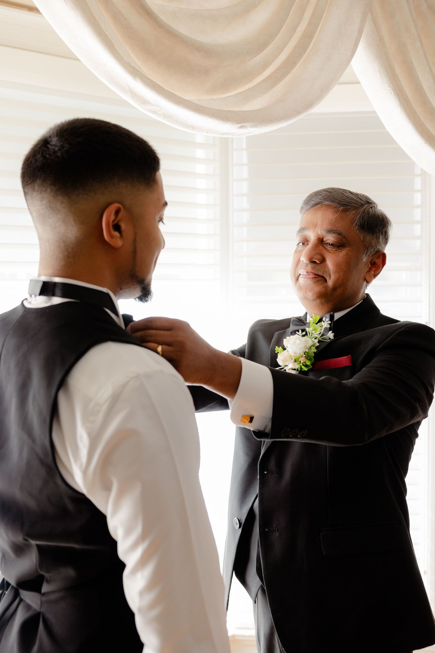 Two people in formal wear help with final wardrobe adjustments near elegant draped windows.