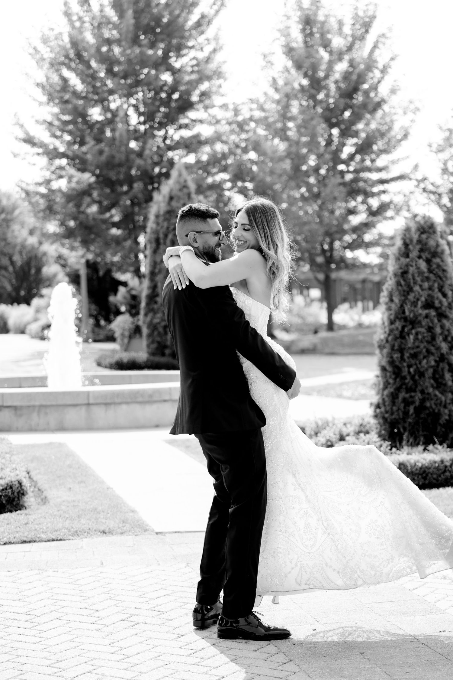 Black and white wedding photo showing joyful couple embracing on brick pathway near garden fountain.