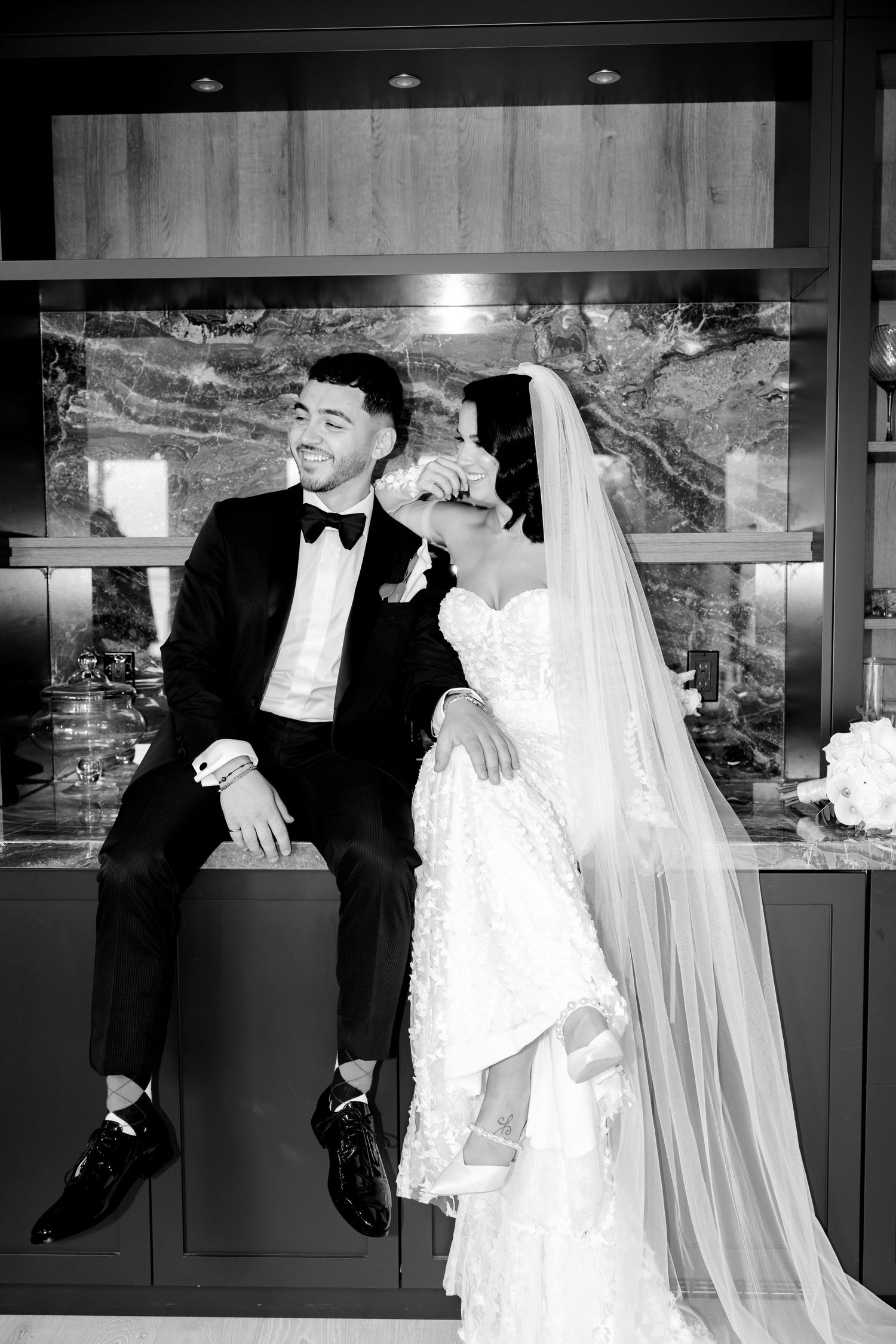 A black and white wedding portrait showing a couple sitting against a modern kitchen backdrop.