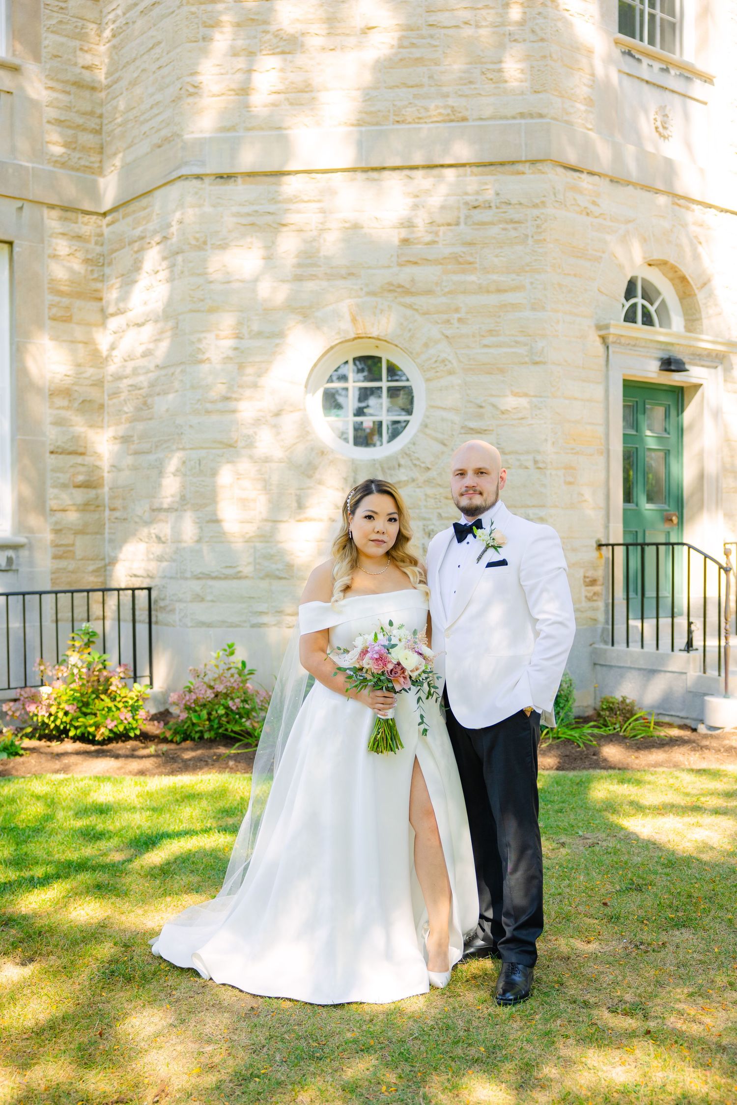 Newlyweds stand together on a sunny day in front of a historic stone church with round window.