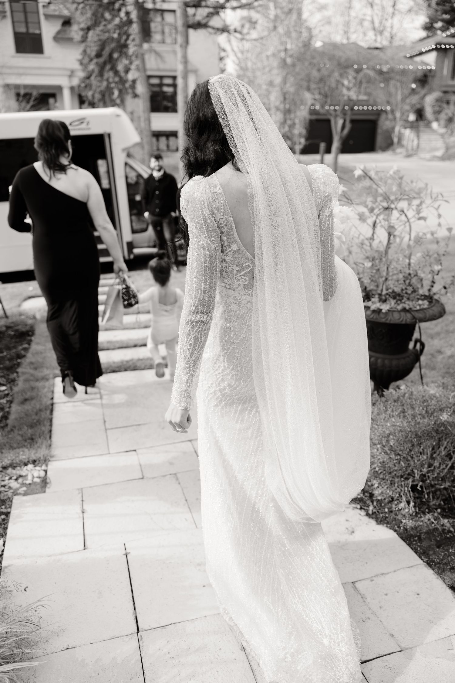 A bride in a long-sleeved lace wedding gown and flowing veil walks along a stone path in black and white.
