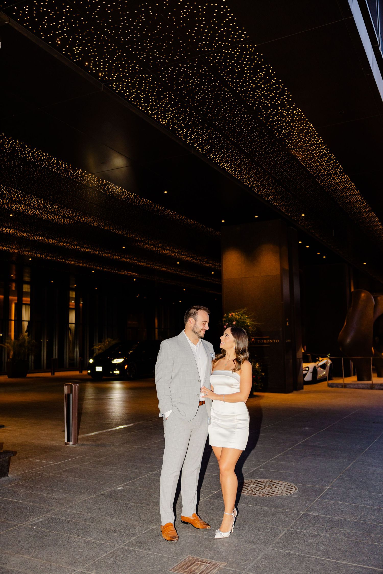 Fashionable pair poses together under decorative lighting in evening setting.