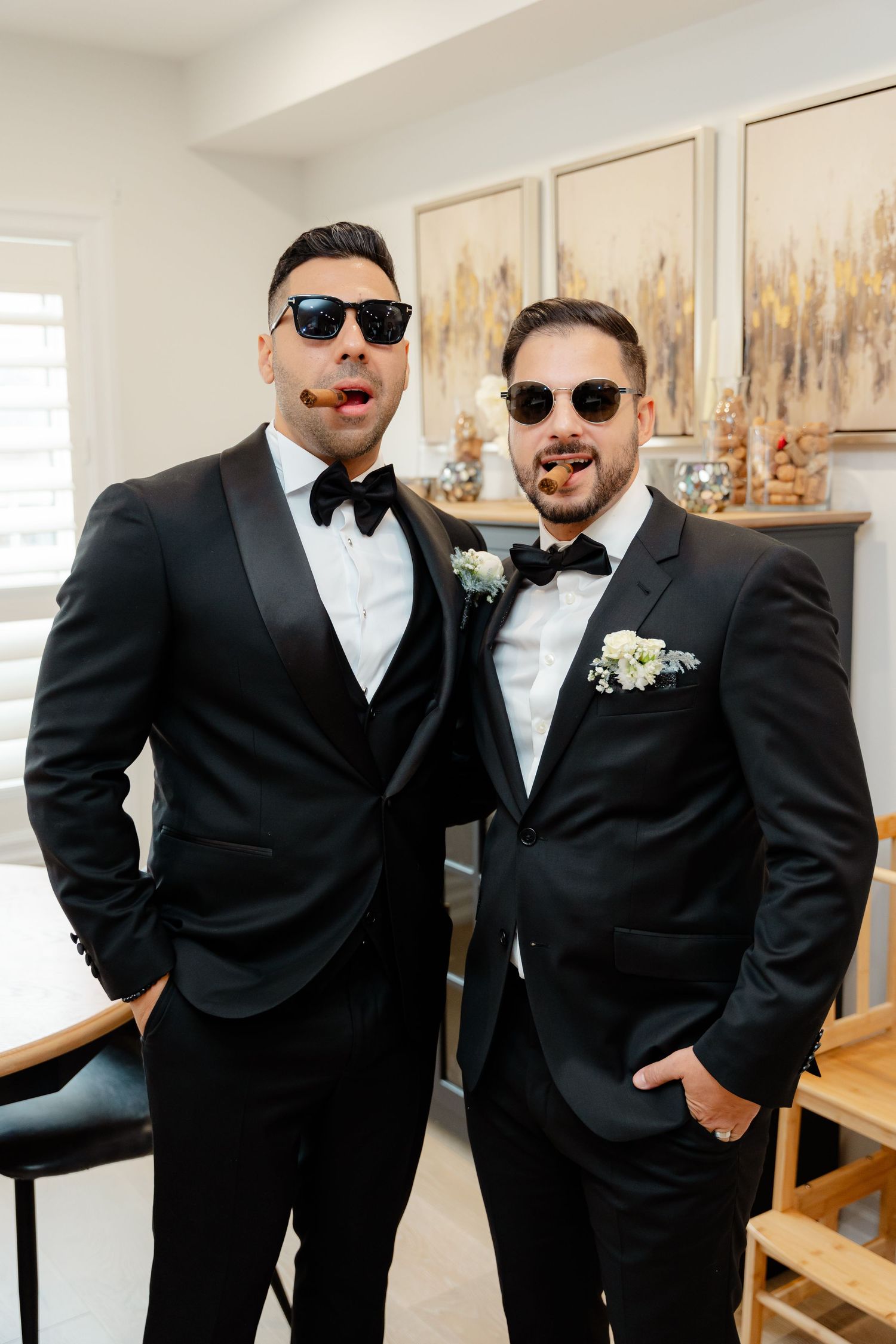 Two groomsmen in black tuxedos and sunglasses pose indoors with art gallery wall decor.
