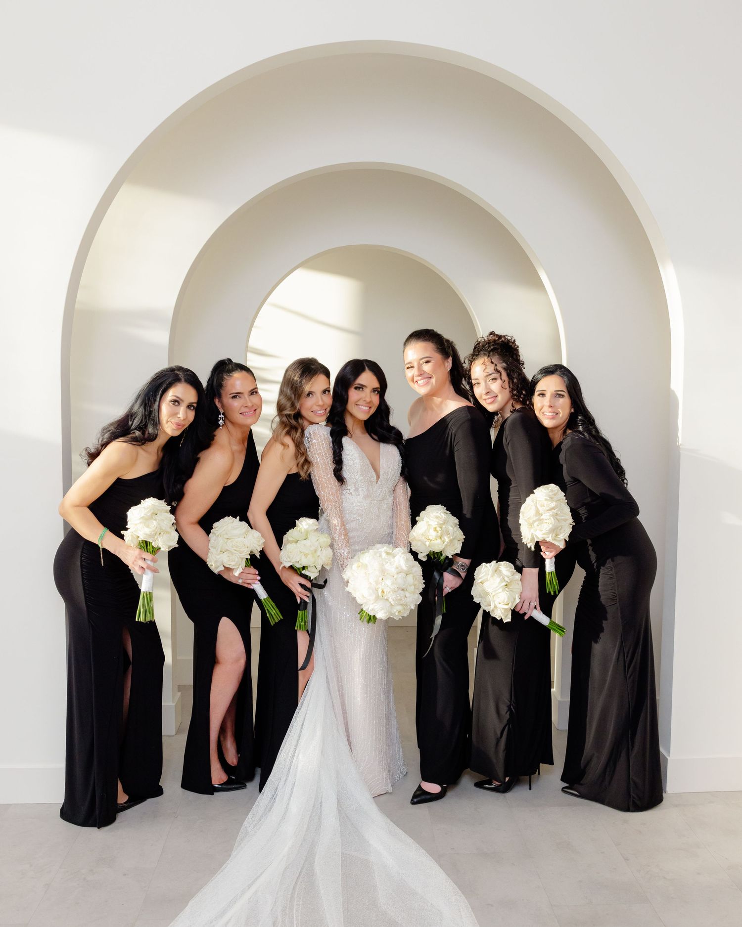 Wedding party portrait under geometric white arches with black gowns and white floral bouquets.