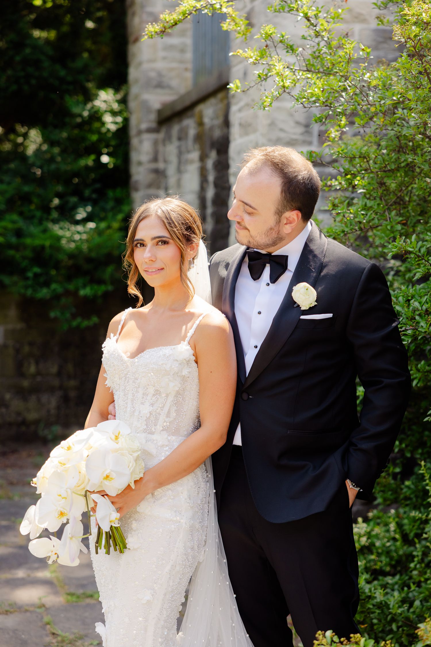 A bride in a lace gown and groom in black tuxedo pose together outdoors near flowering foliage on their wedding day.