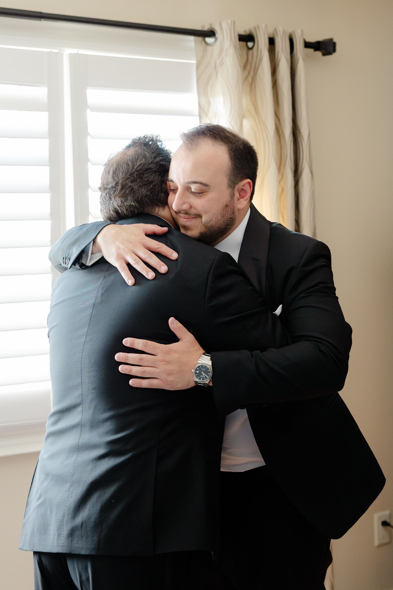 Two people share an emotional embrace while wearing formal attire in a room with natural lighting and curtains.