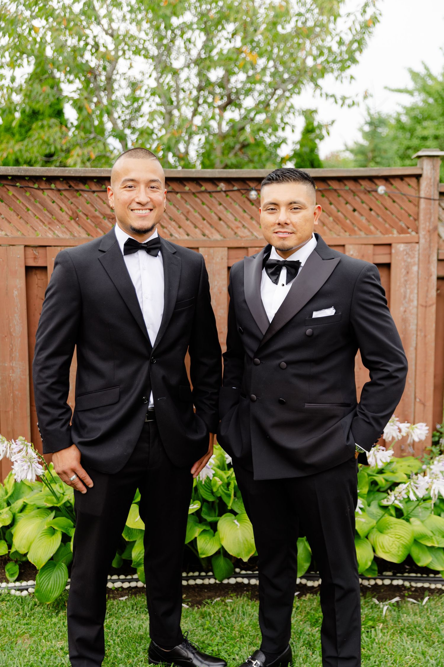 A sequence of groomsmen in black tuxedos posed against a wooden fence and garden backdrop.