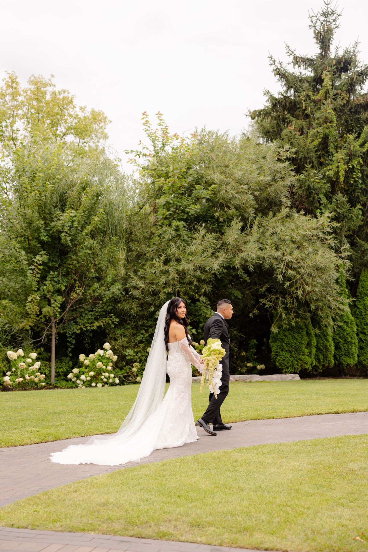 A couple walks along a garden path surrounded by lush greenery and white hydrangeas on their wedding day.