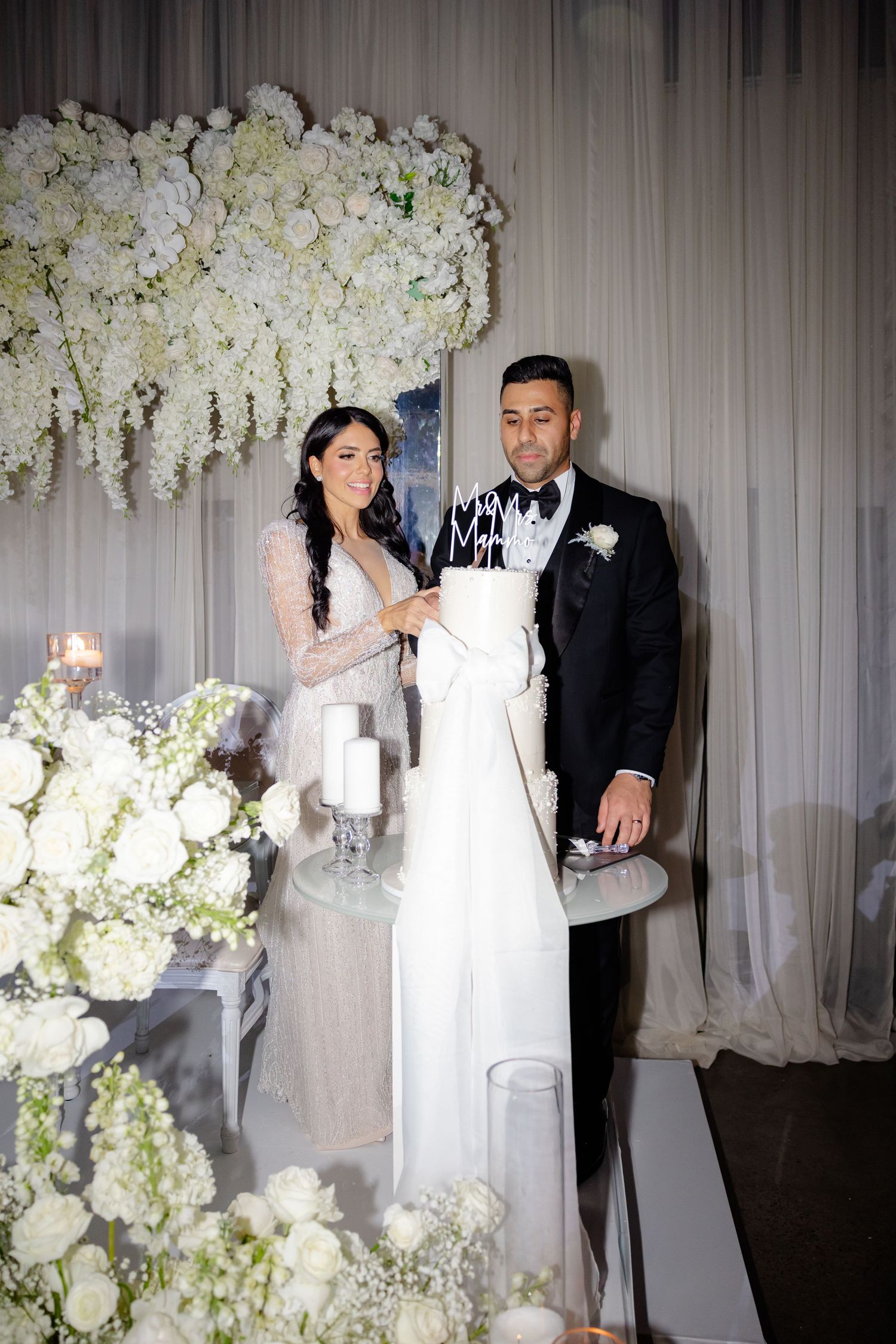 Bride and groom stand with their tall white wedding cake at elegant reception.