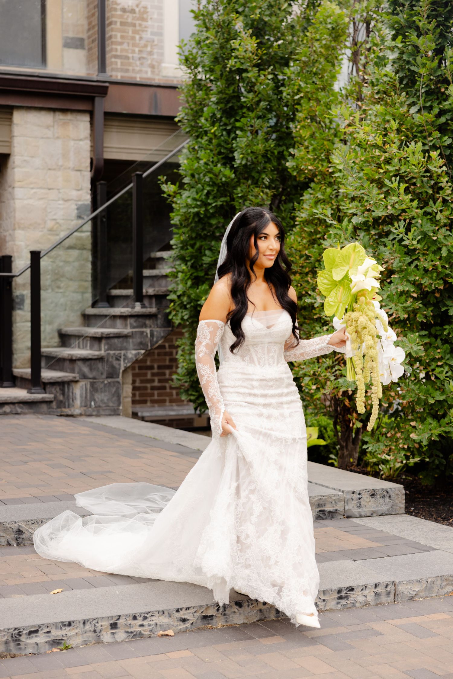 A person in a flowing white lace wedding dress descends stone steps in a garden setting.