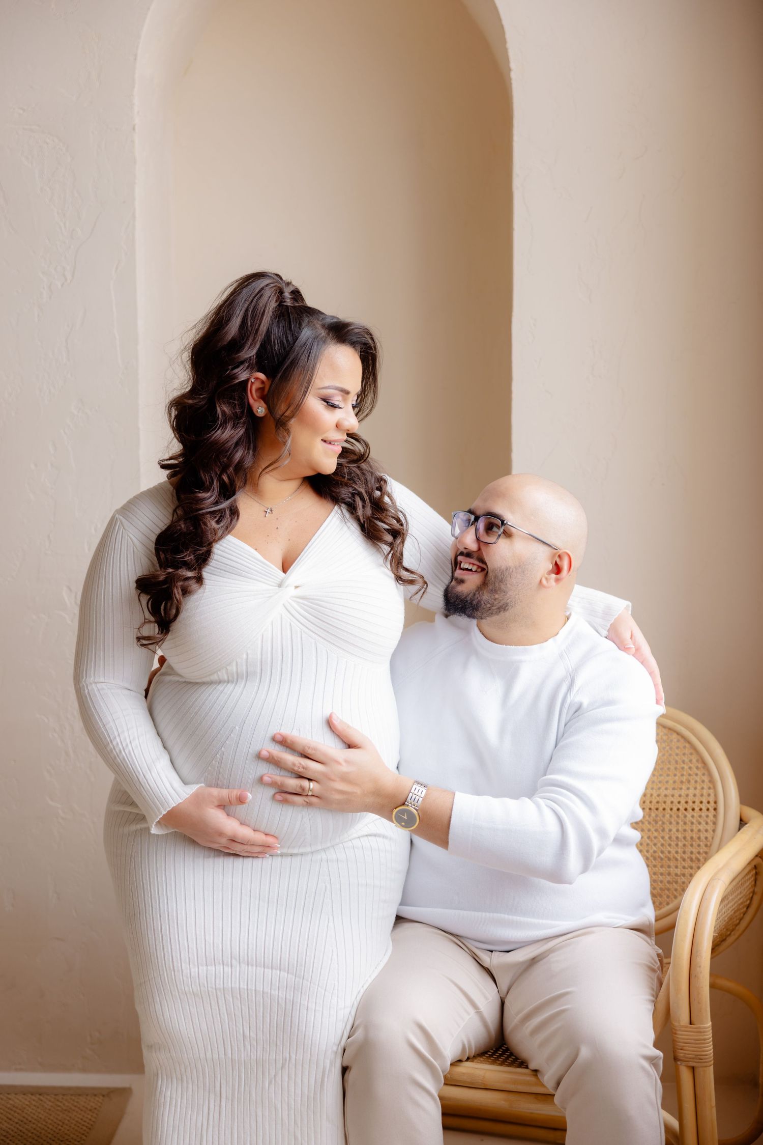 Expectant couple in matching white outfits share tender moment in front of arched wall during maternity photoshoot.