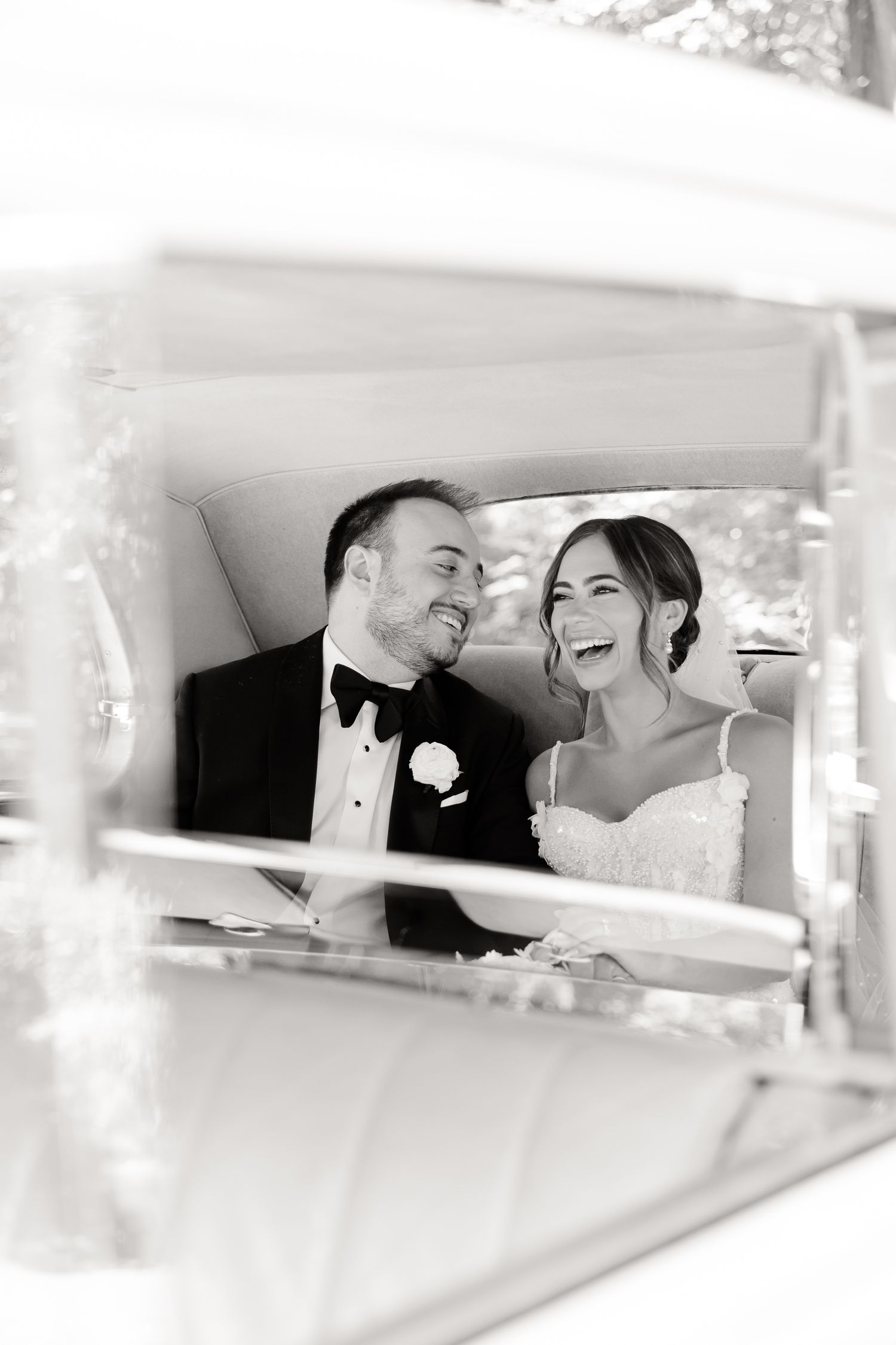 A joyful black and white wedding photograph showing a couple laughing together while seated in a vintage car.