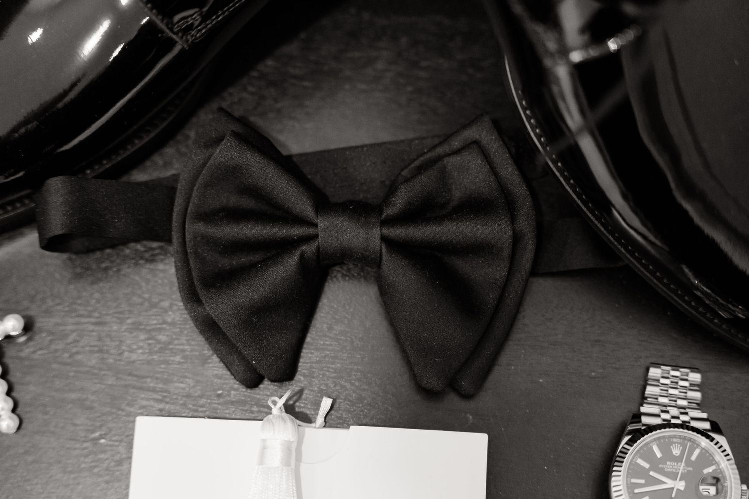 Black bow tie and watch arranged with a white tag in black and white photography.
