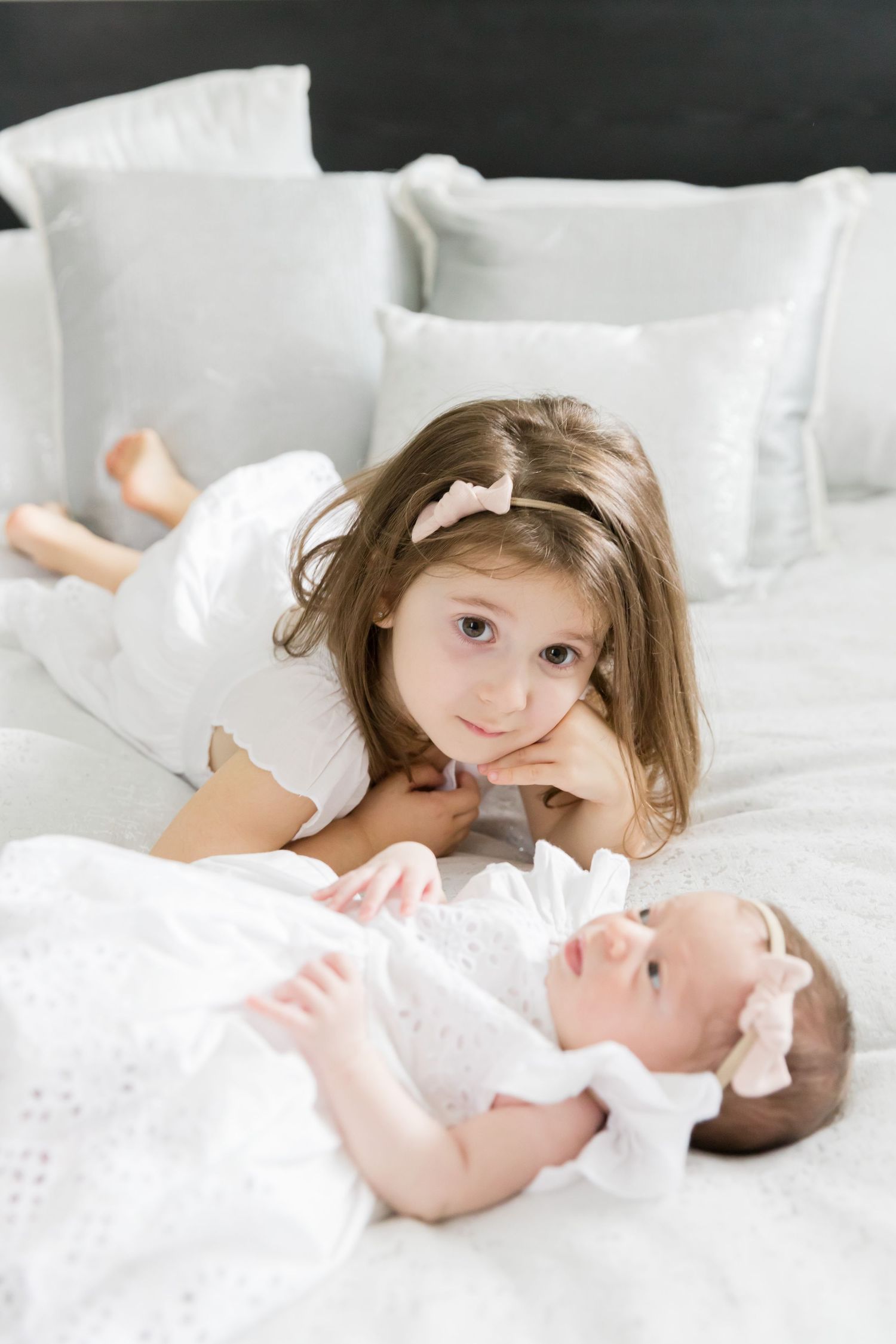 Two young children in white lying on bed with white pillows and bedding.