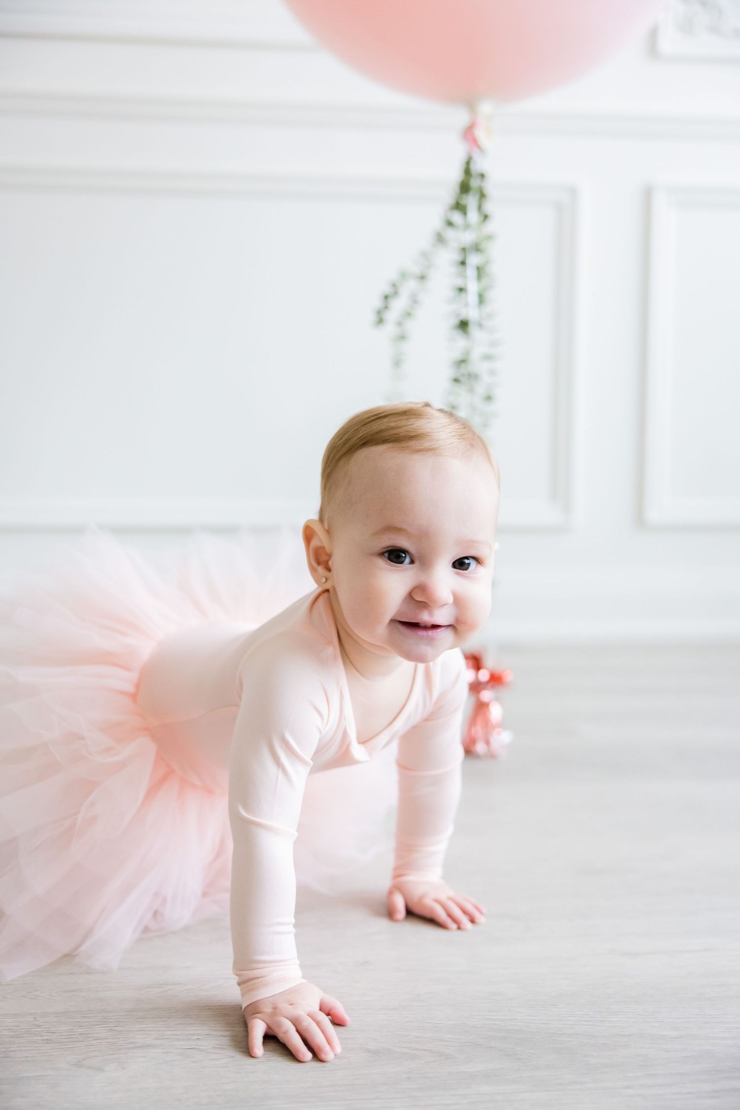 Toddler in pink ballet outfit crawls on floor with large balloon and greenery in background.