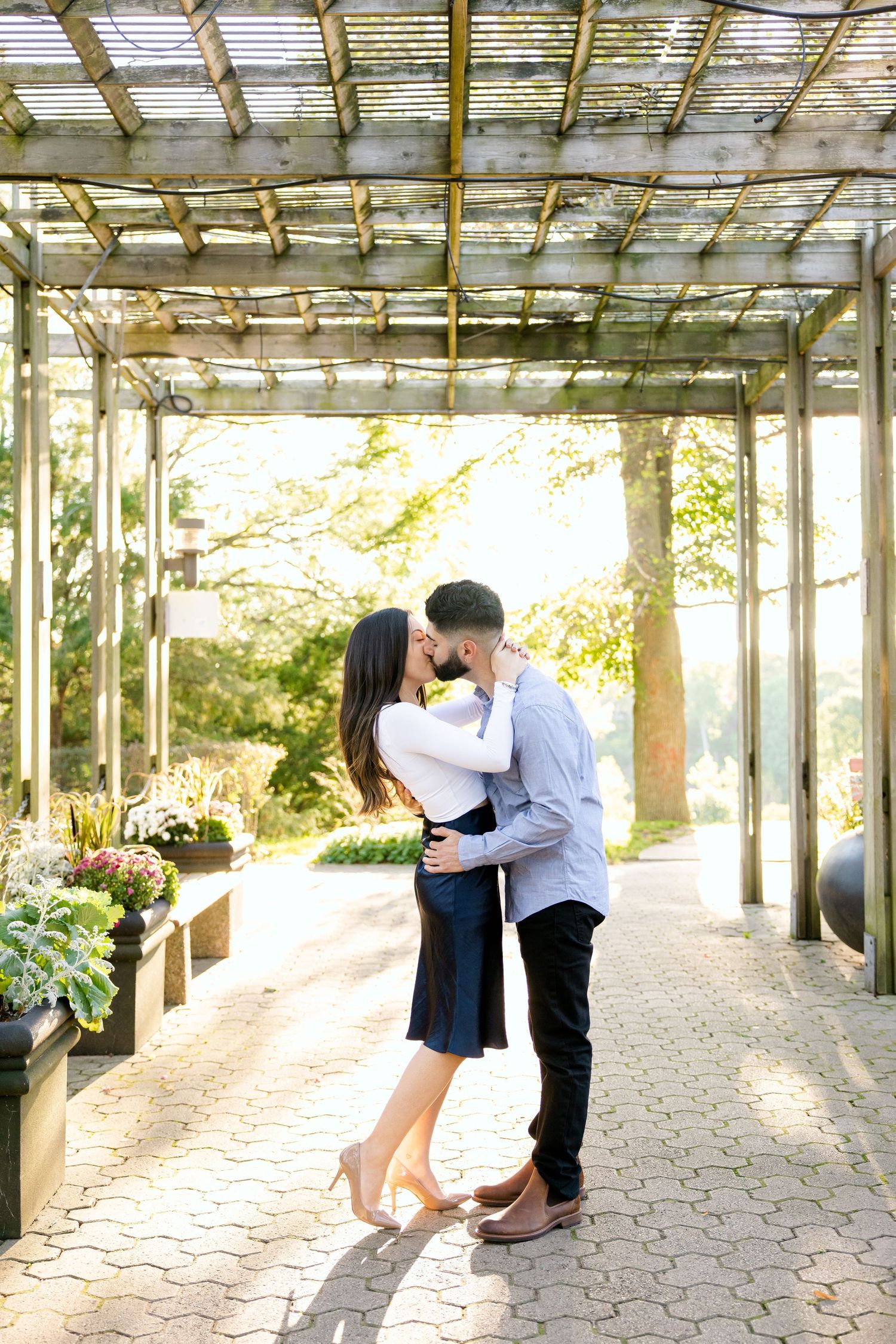 A couple sharing a romantic moment under a sunlit pergola surrounded by garden planters.