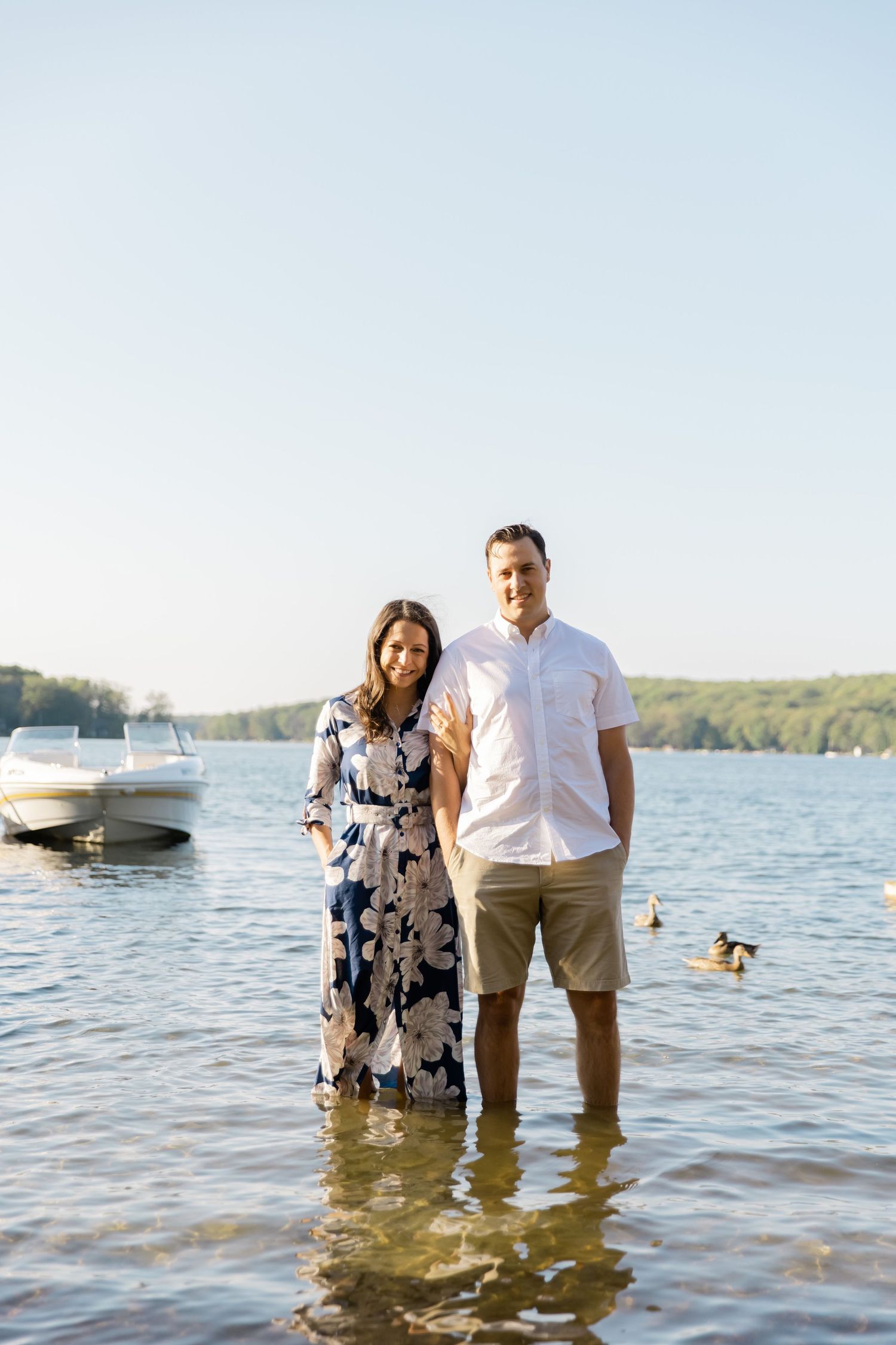 A couple stands together in ankle-deep lake water with boats and trees in the background.