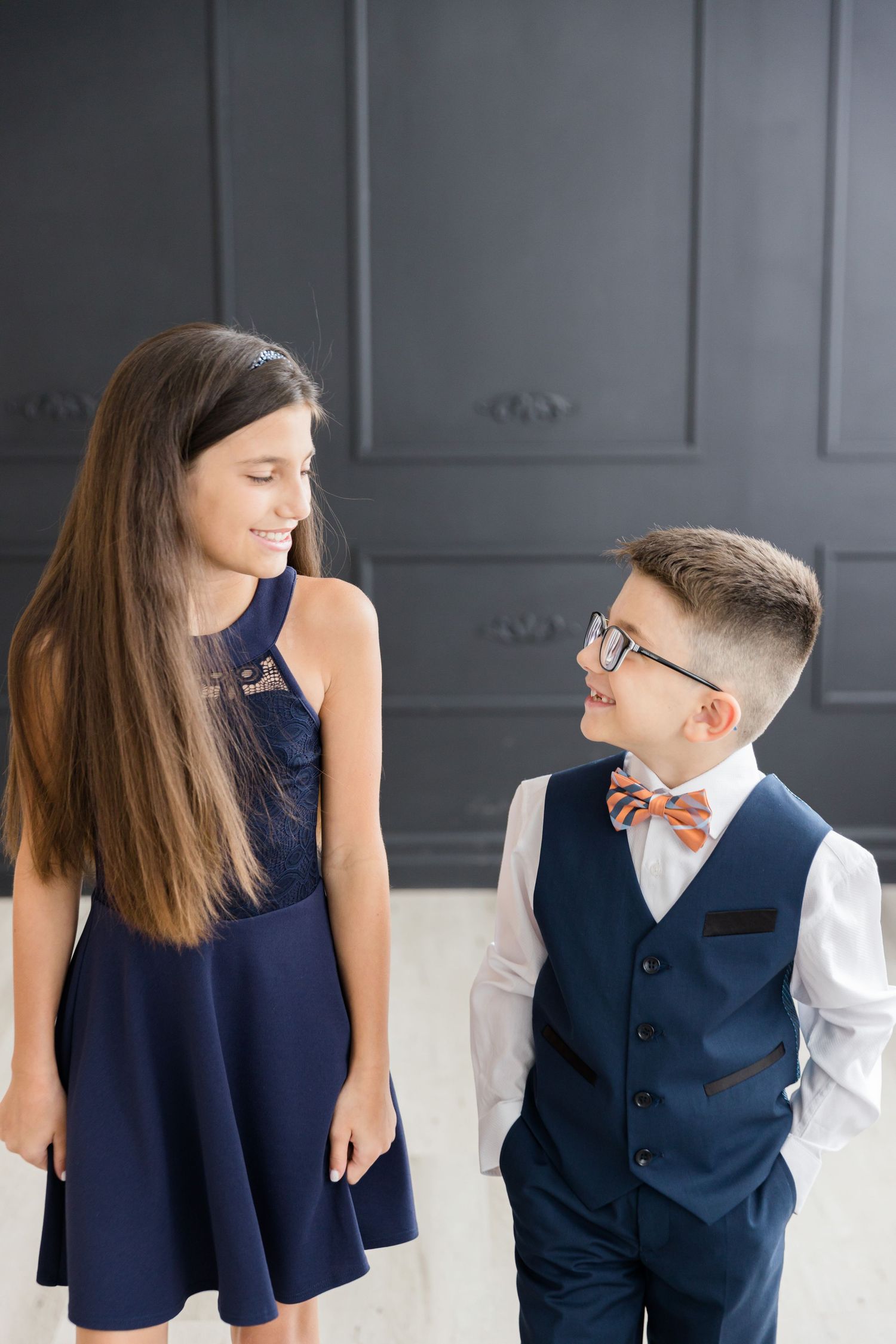 Brother and sister in coordinating navy outfits share a candid moment against dark wall panels.