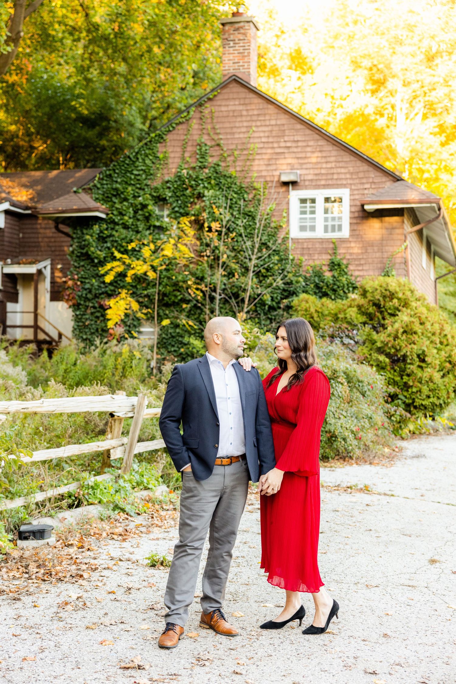 Couple in formal attire stand together near rustic brown house with fall foliage.