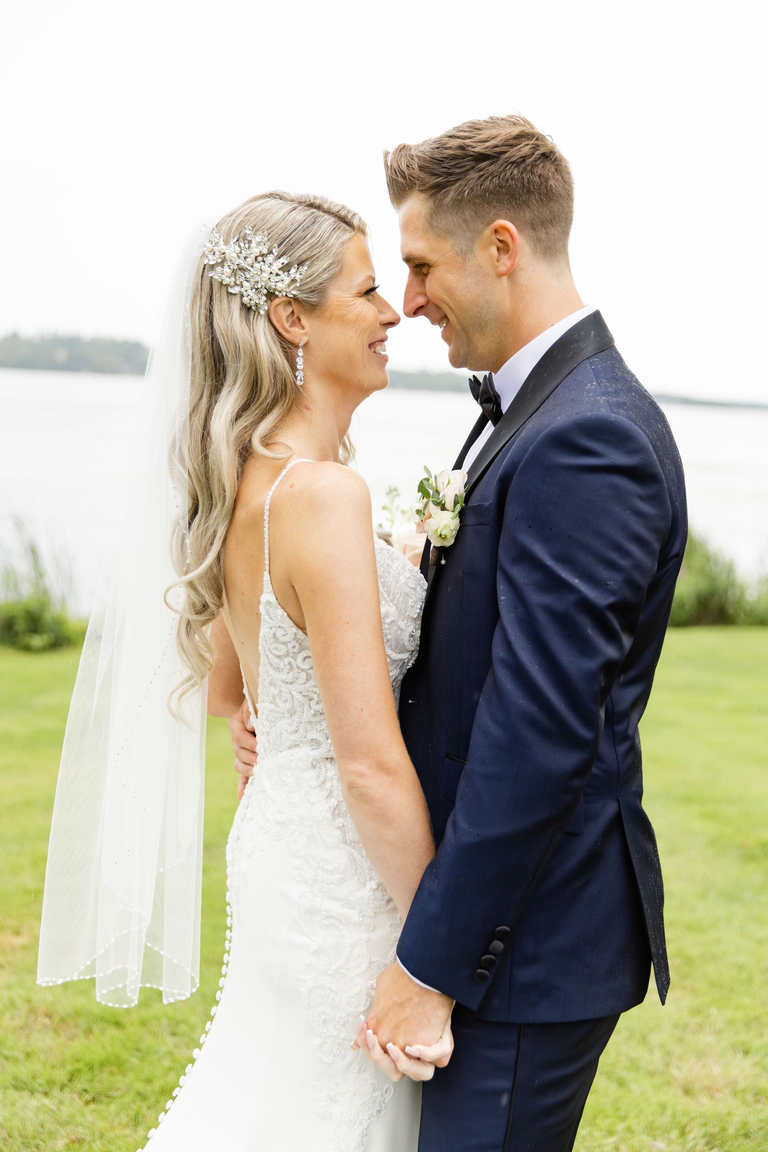 Intimate moment captured between couple in navy suit and lace wedding dress against waterfront backdrop.