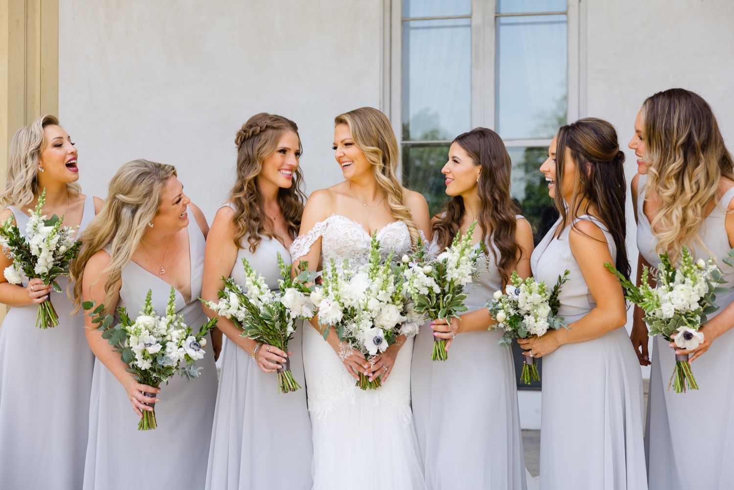 Candid moment of bridal party in soft gray dresses with white and green bouquets.