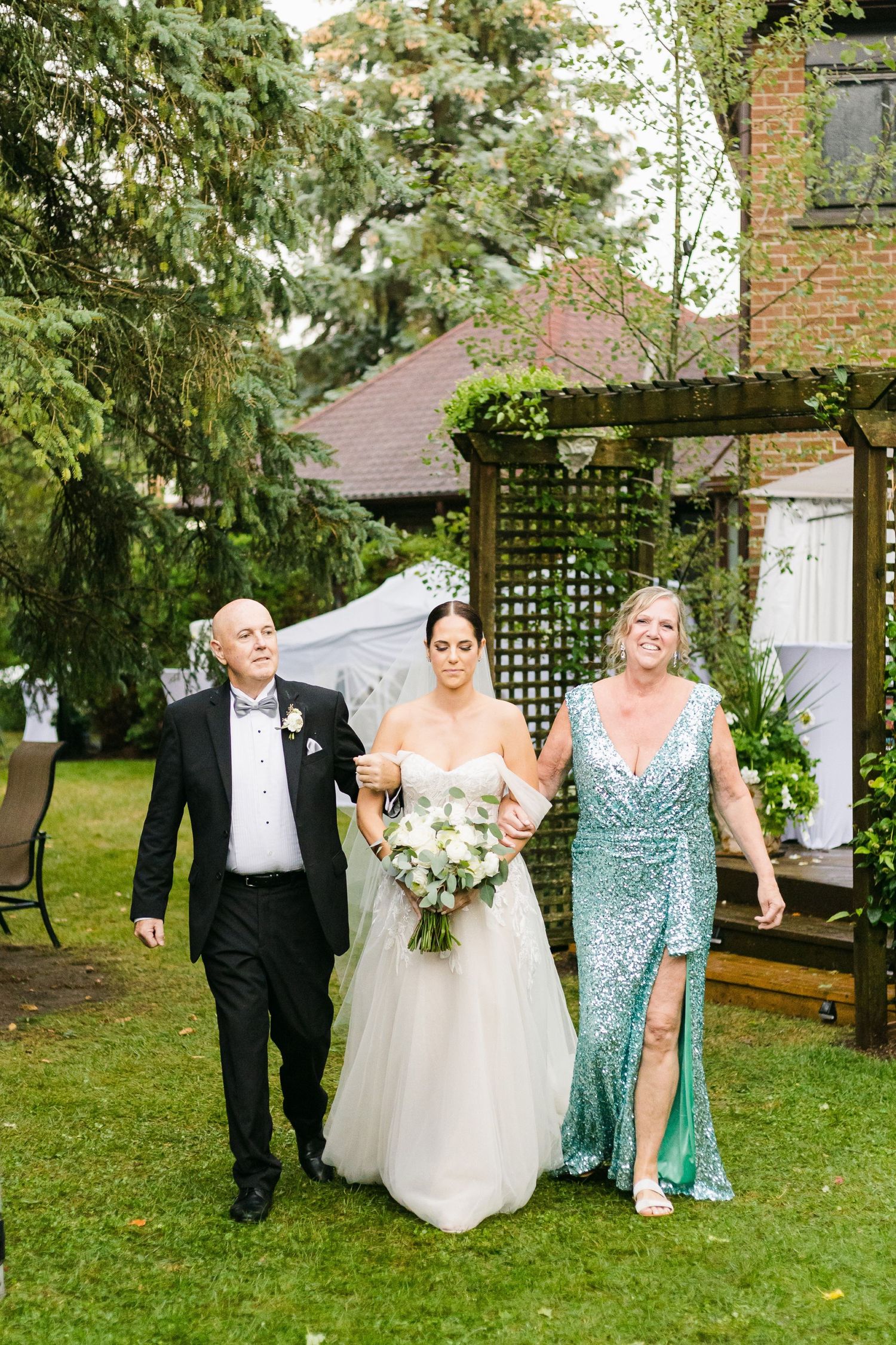 Formal couple walks with someone in an elegant teal gown through a garden setting with a pergola.