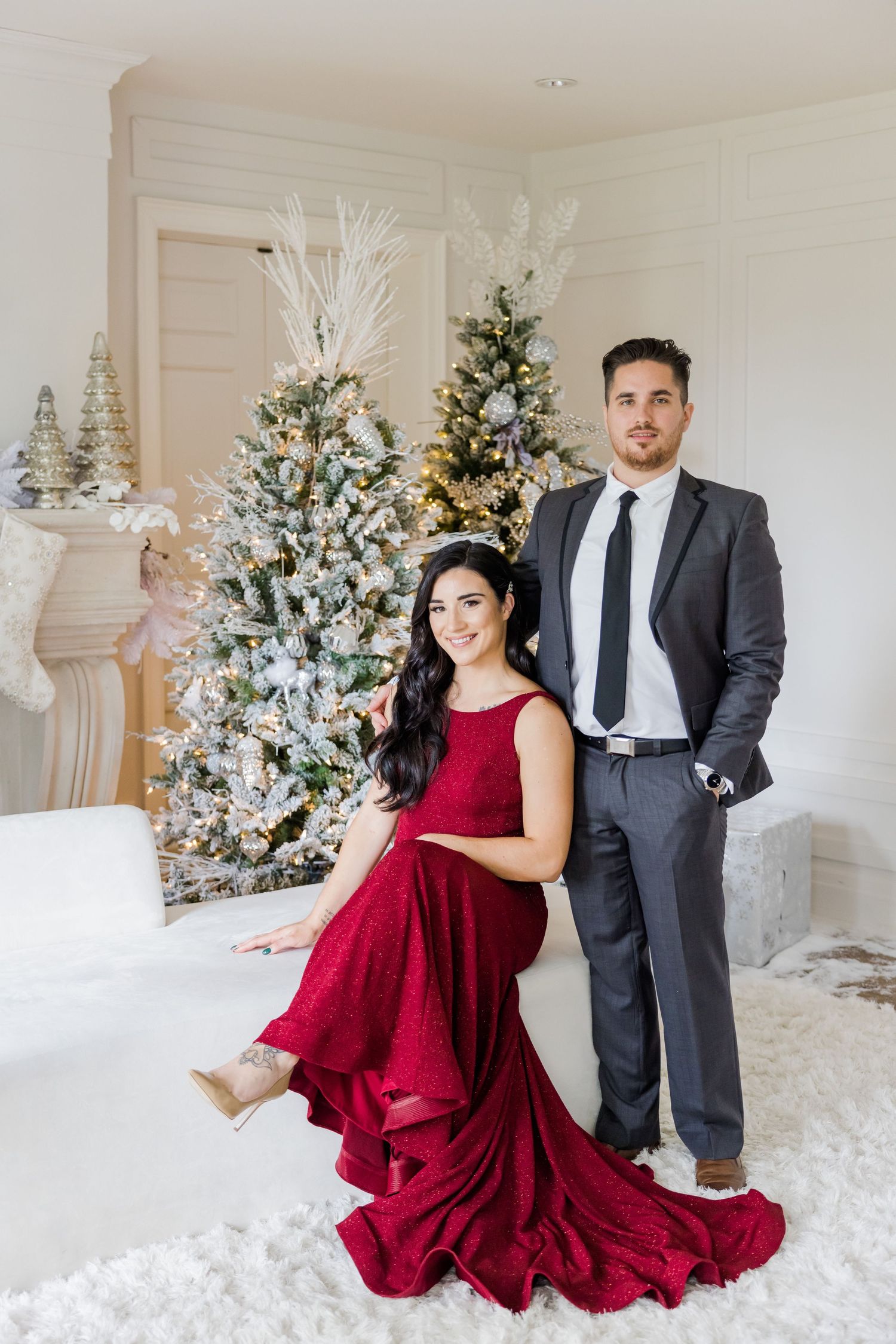 Formal Christmas portrait with dramatic red gown and grey suit among decorated trees.