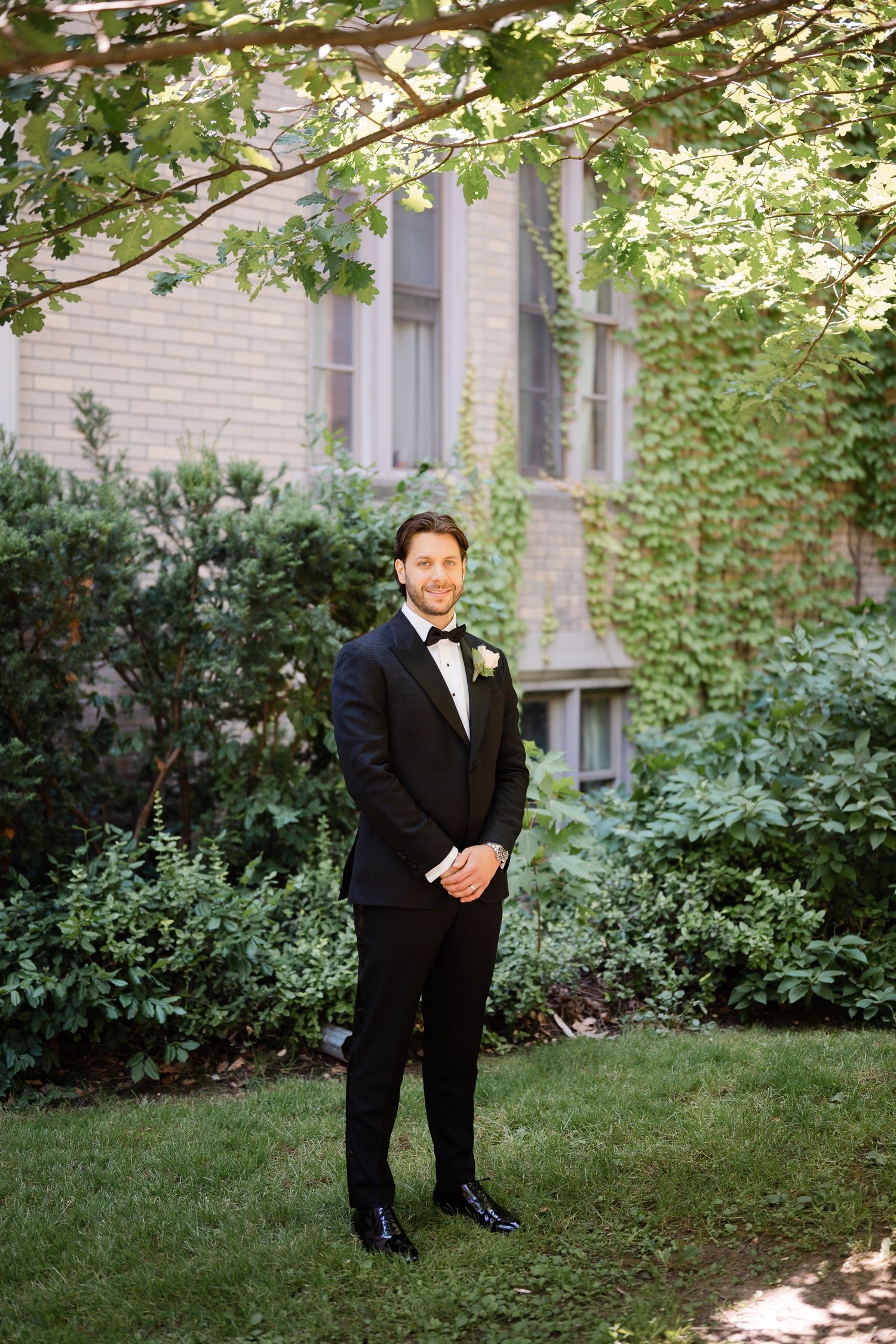 Groom in classic black tuxedo poses in garden setting with dappled sunlight.