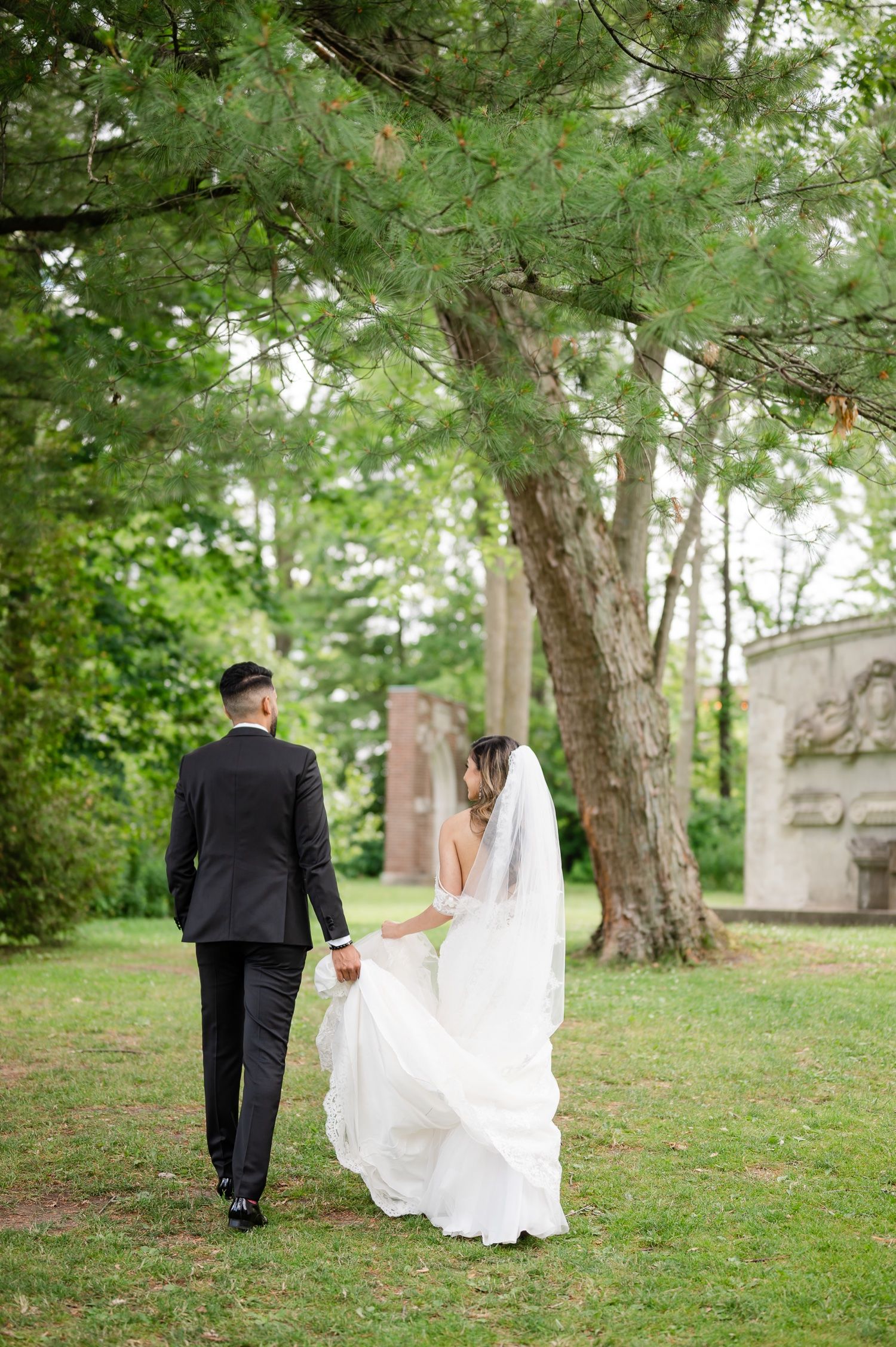 A couple in wedding attire strolls through a lush garden with historic architecture in background.