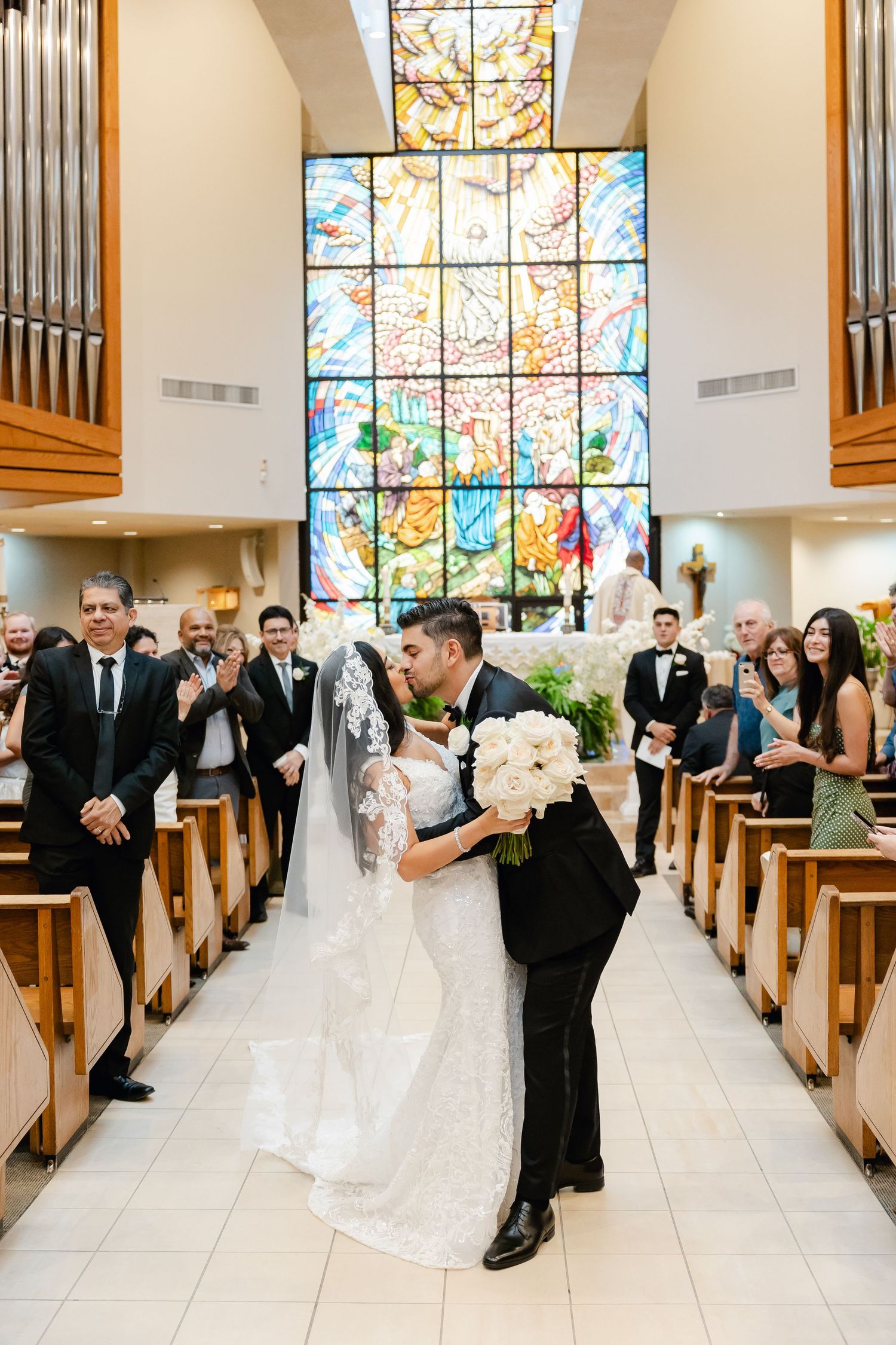 Church wedding ceremony with guests watching couple share first kiss under stained glass window.