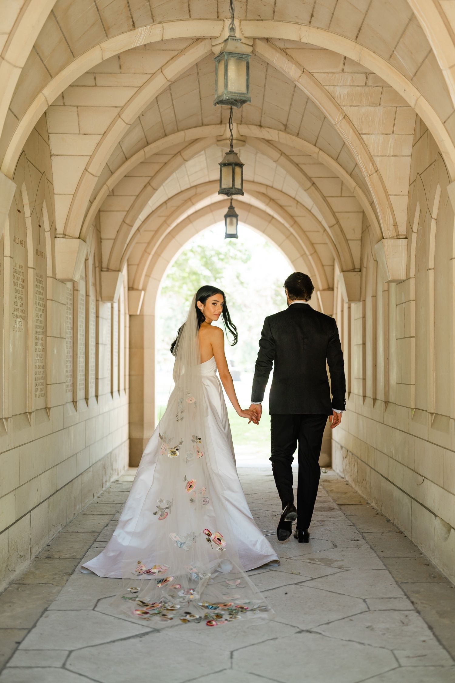 Bride in floral train wedding dress walks with groom through elegant stone archway.