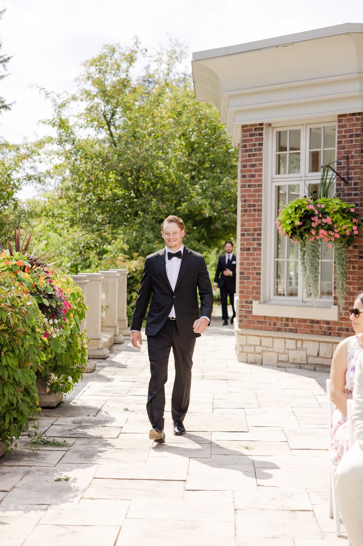 Groom in black bow tie and tuxedo walking outside brick building on sunny day.