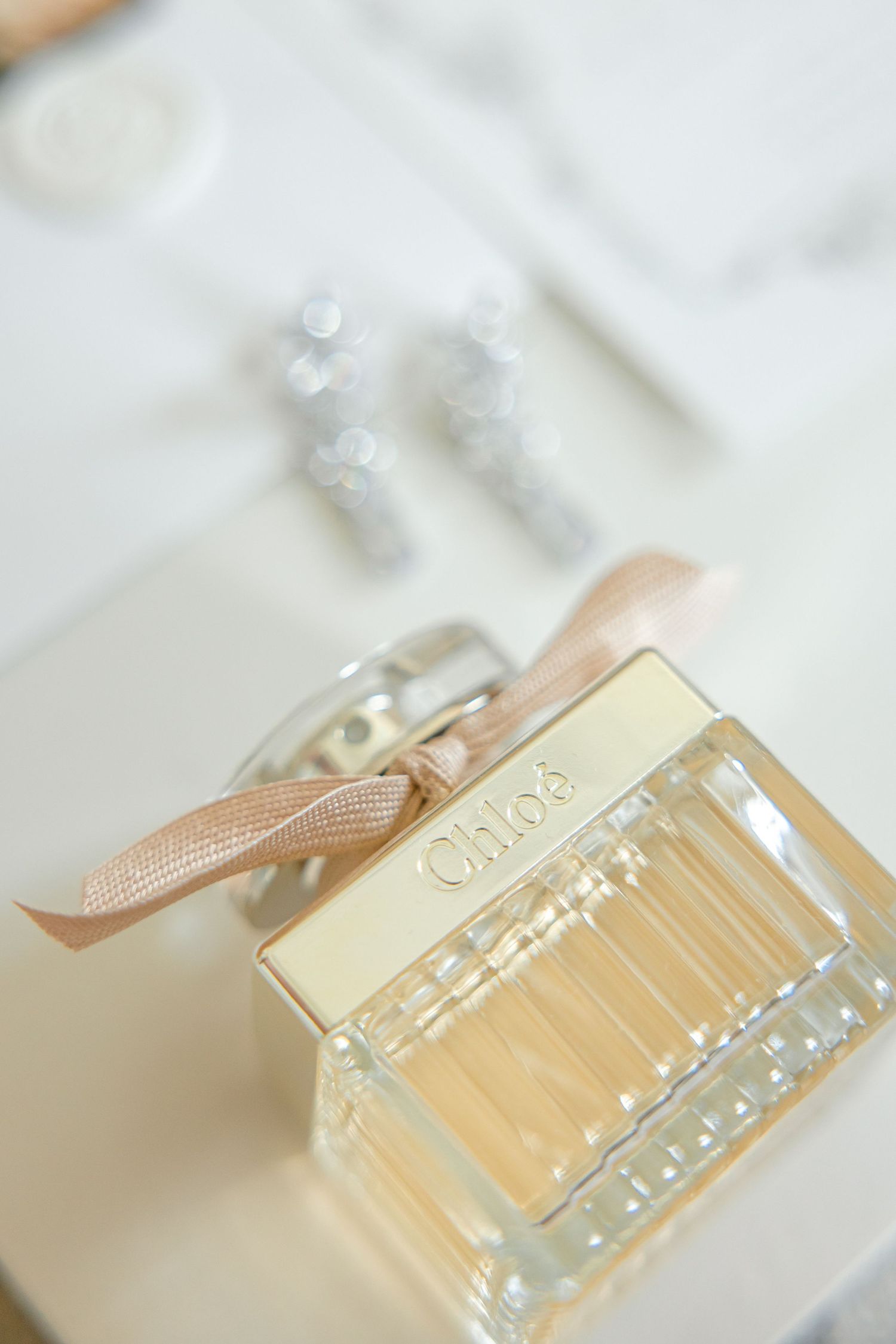 Luxury perfume bottle with peach ribbon on white surface next to crystal earrings.