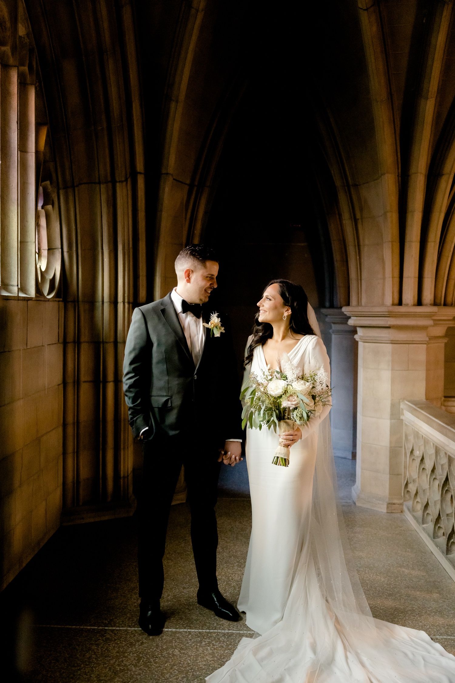 Wedding couple gazes at each other in a dramatic stone hallway with gothic arches and natural light.