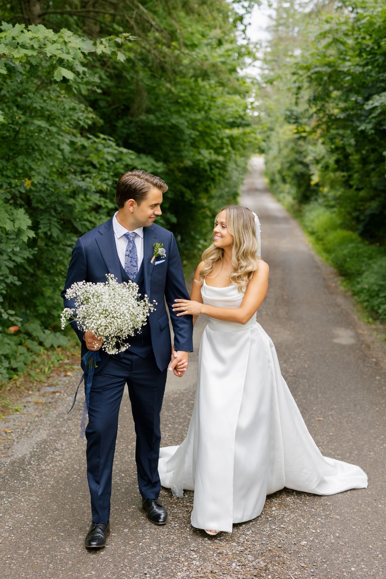 Couple walks along forest path as groom carries baby's breath bouquet.