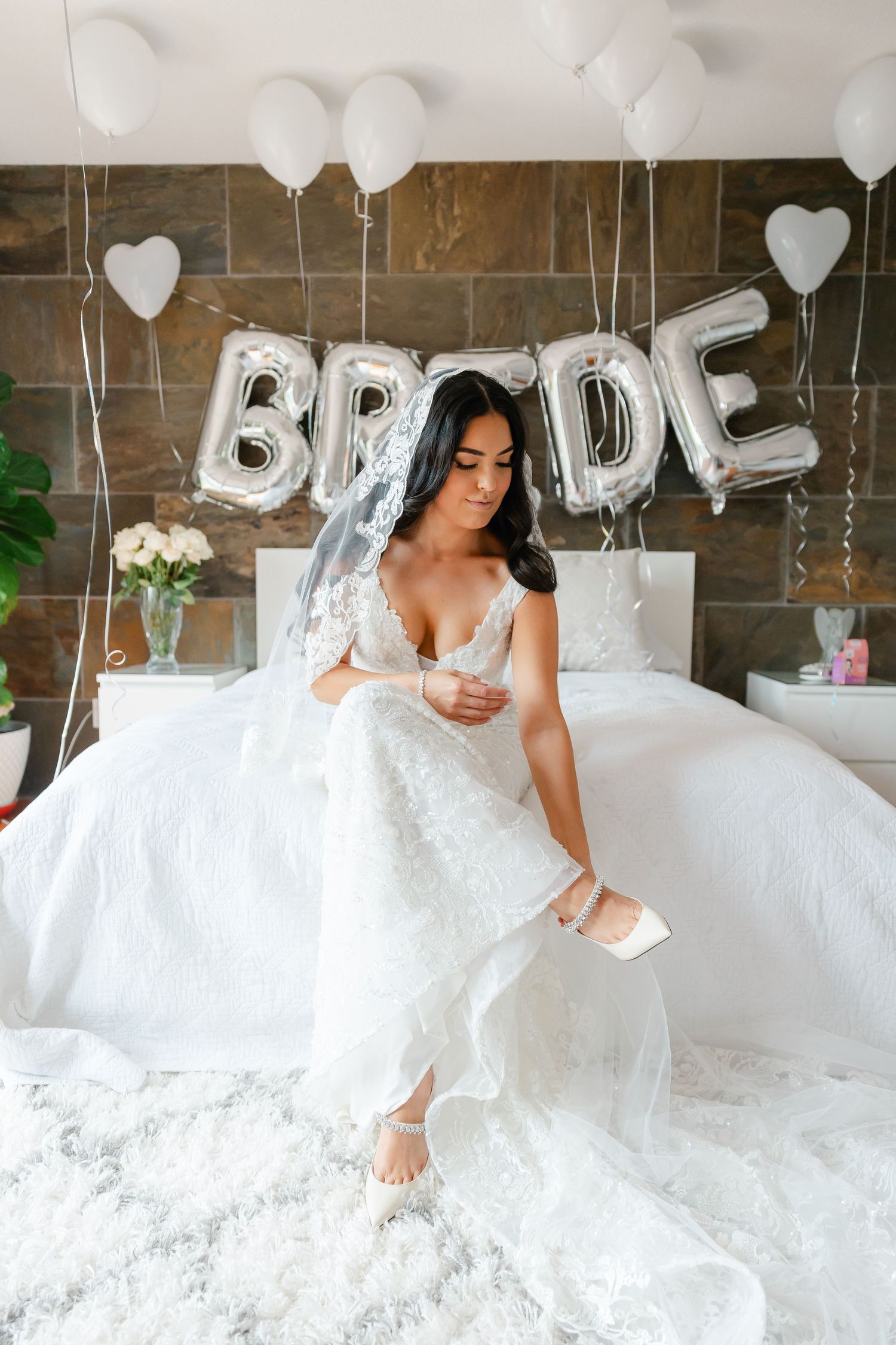 Bride in white gown sits on bed with BRIDE balloon letters and heart decorations.