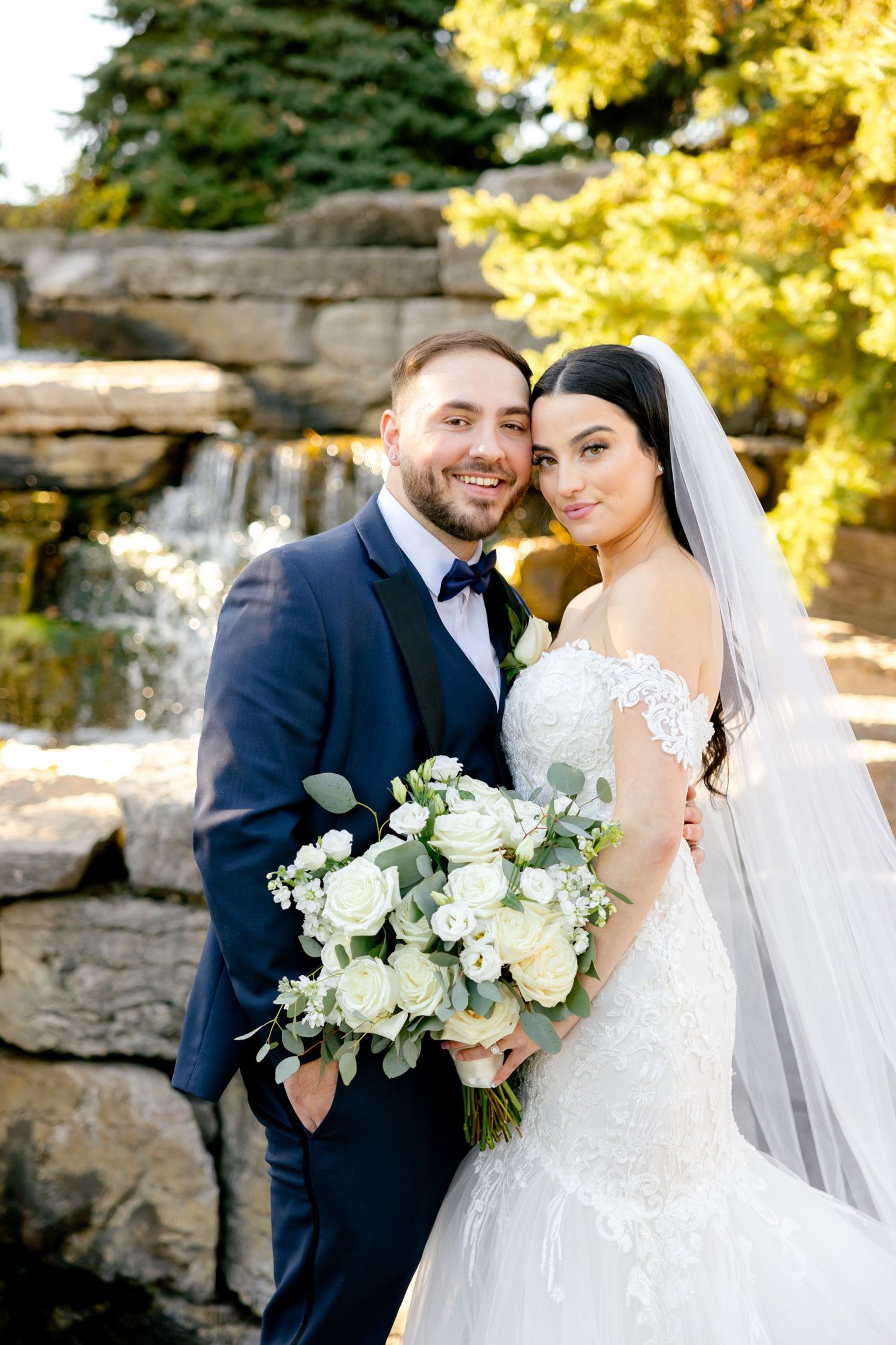 A wedding couple poses in front of a stone waterfall feature surrounded by autumn foliage.