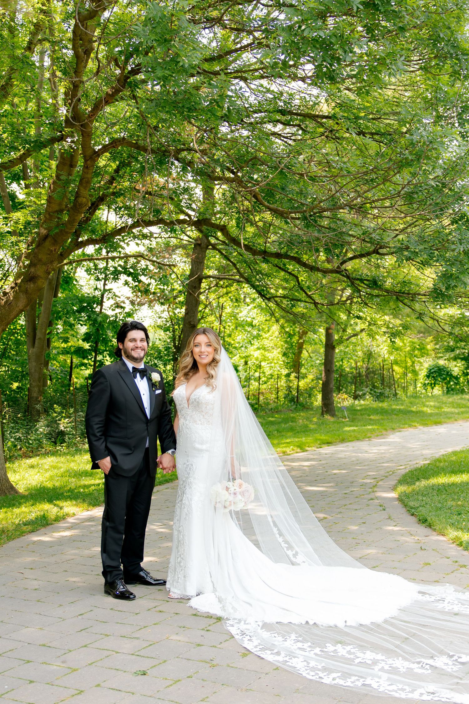 A bride and groom stand together on a paved path with a flowing cathedral veil and green forest backdrop.